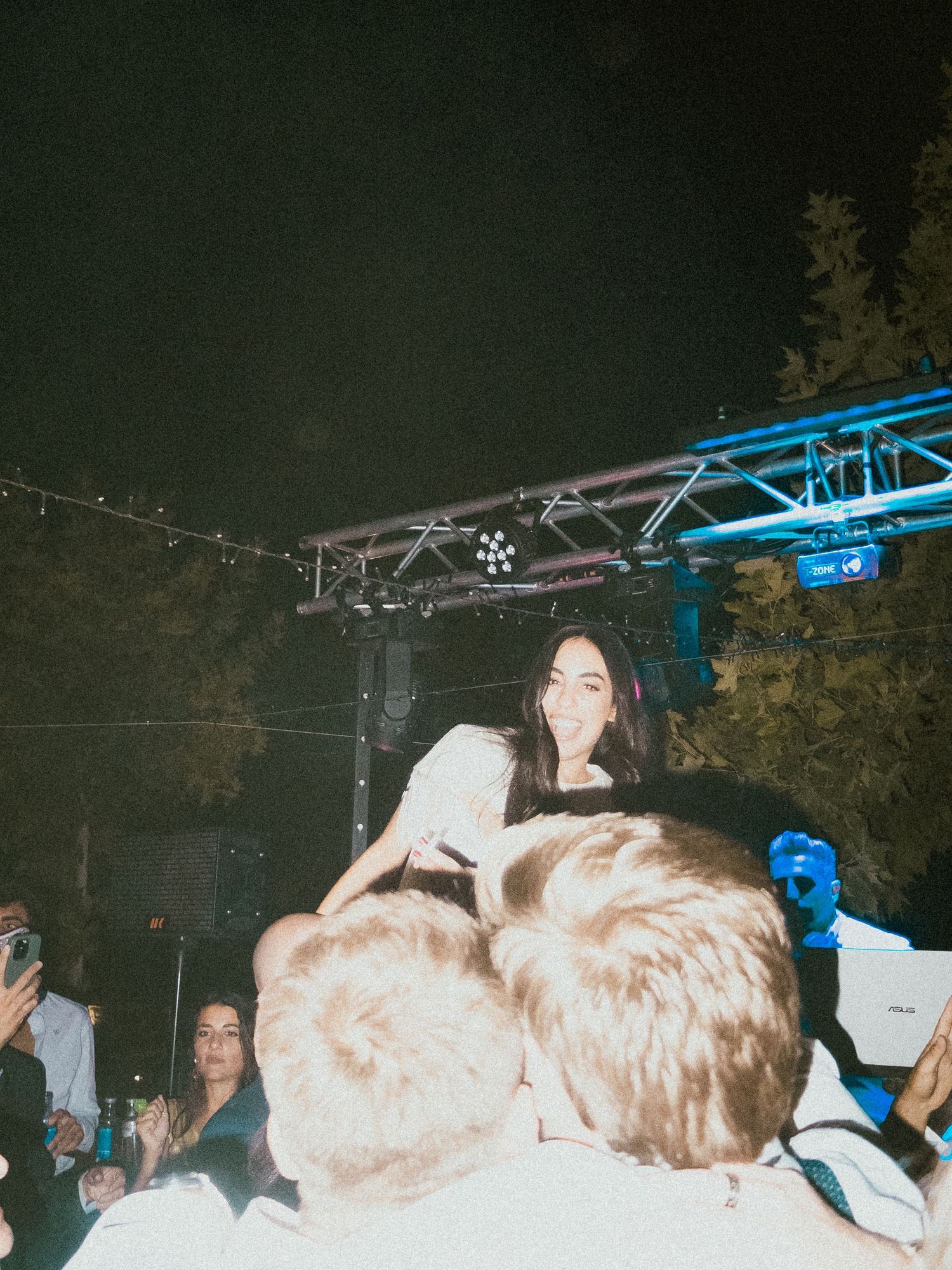 Nighttime outdoor party scene with lighting rigs and dancing crowd.