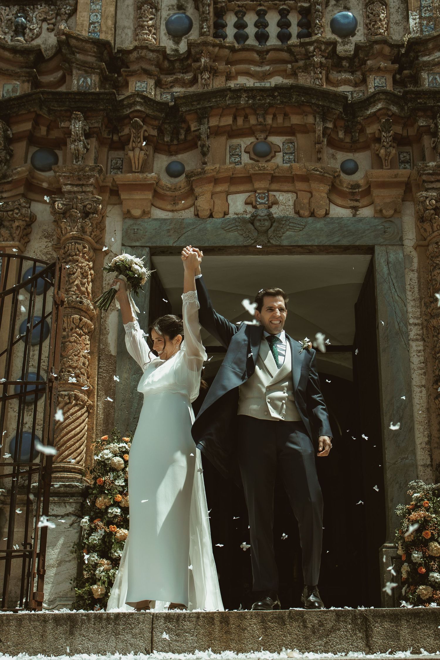 Wedding couple exits ornate stone church entrance while guests throw flower petals in celebration.