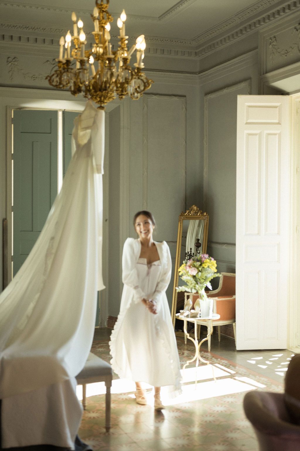 Sunlit room with flowing white wedding dress and chandelier creating an ethereal atmosphere.