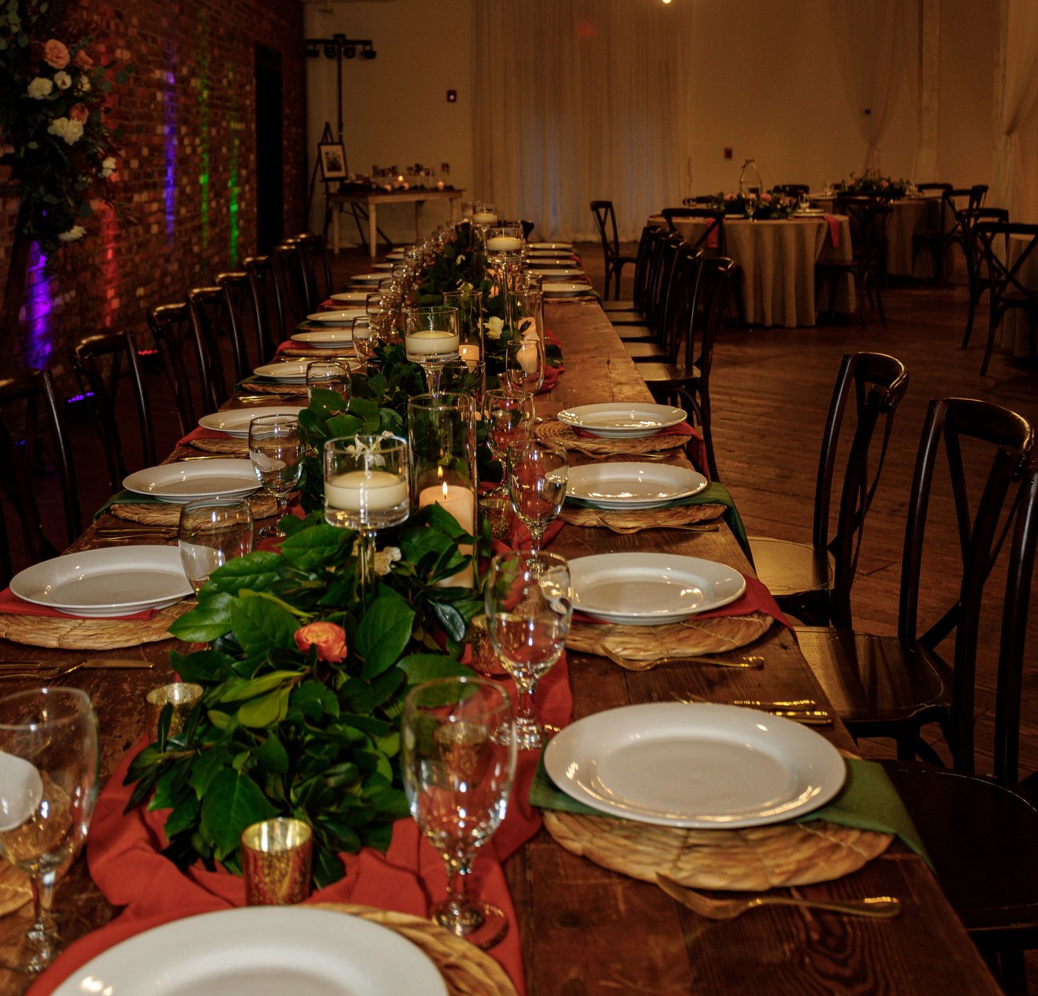 Elegant wedding reception table setting with white plates, red tablecloth and greenery centerpieces in a rustic venue.
