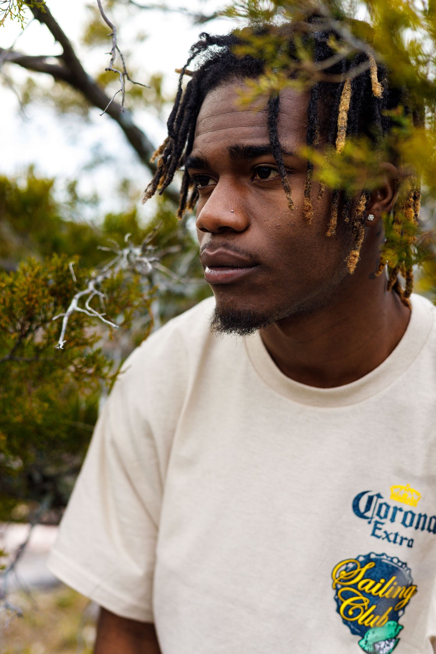 Person wearing a white Corona t-shirt poses against a blurred outdoor background with trees.