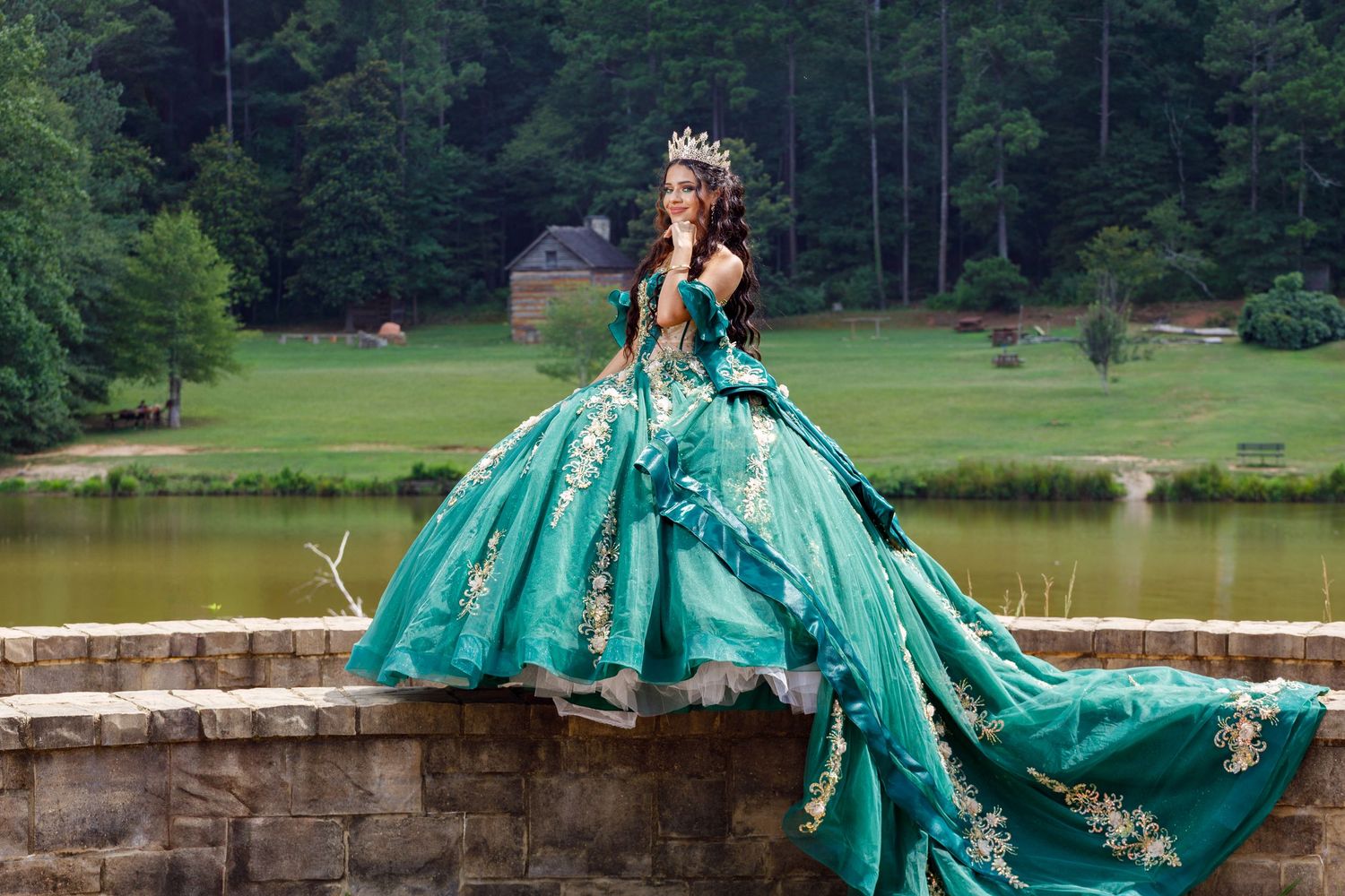 A stunning turquoise quinceañera ball gown with gold embellishments sparkles by a serene lake with forest backdrop.