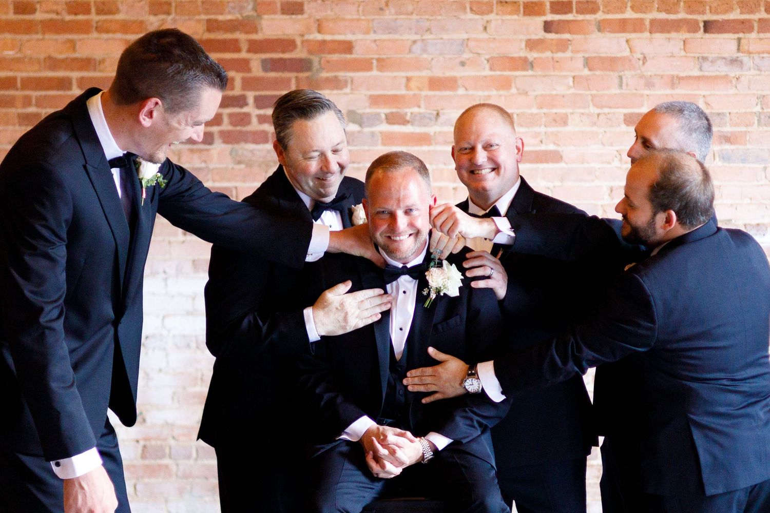 Groomsmen in dark suits sharing a playful moment against a brick wall backdrop.