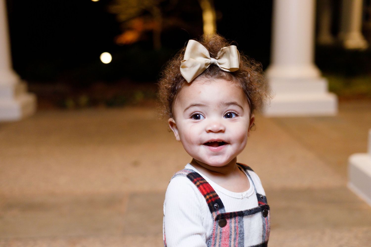 A baby wearing a red and white striped outfit smiles while sitting on a carpeted floor.