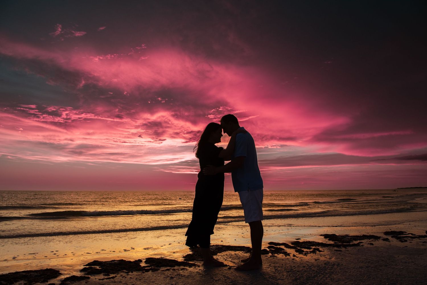 Silhouettes of figures stand together against a dramatic pink and purple sunset sky over a beach shoreline.