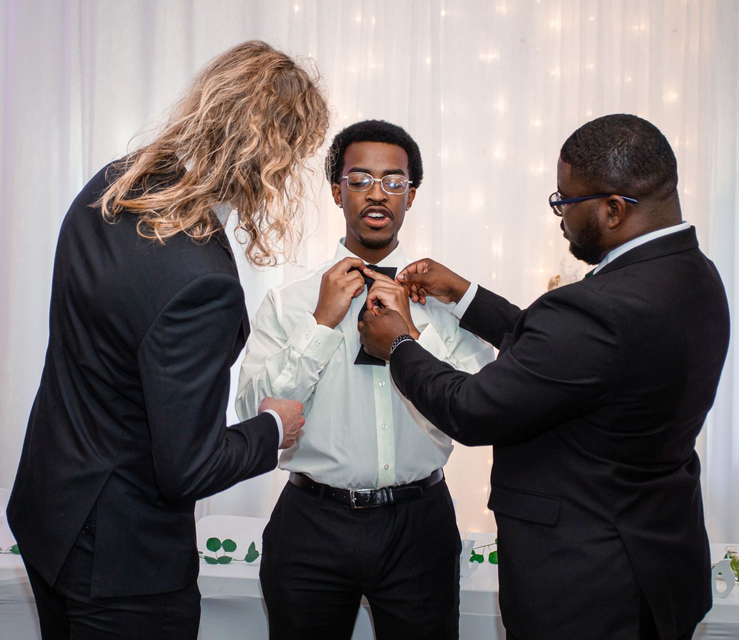Two people helping adjust a bowtie on a guest at a formal wedding event.