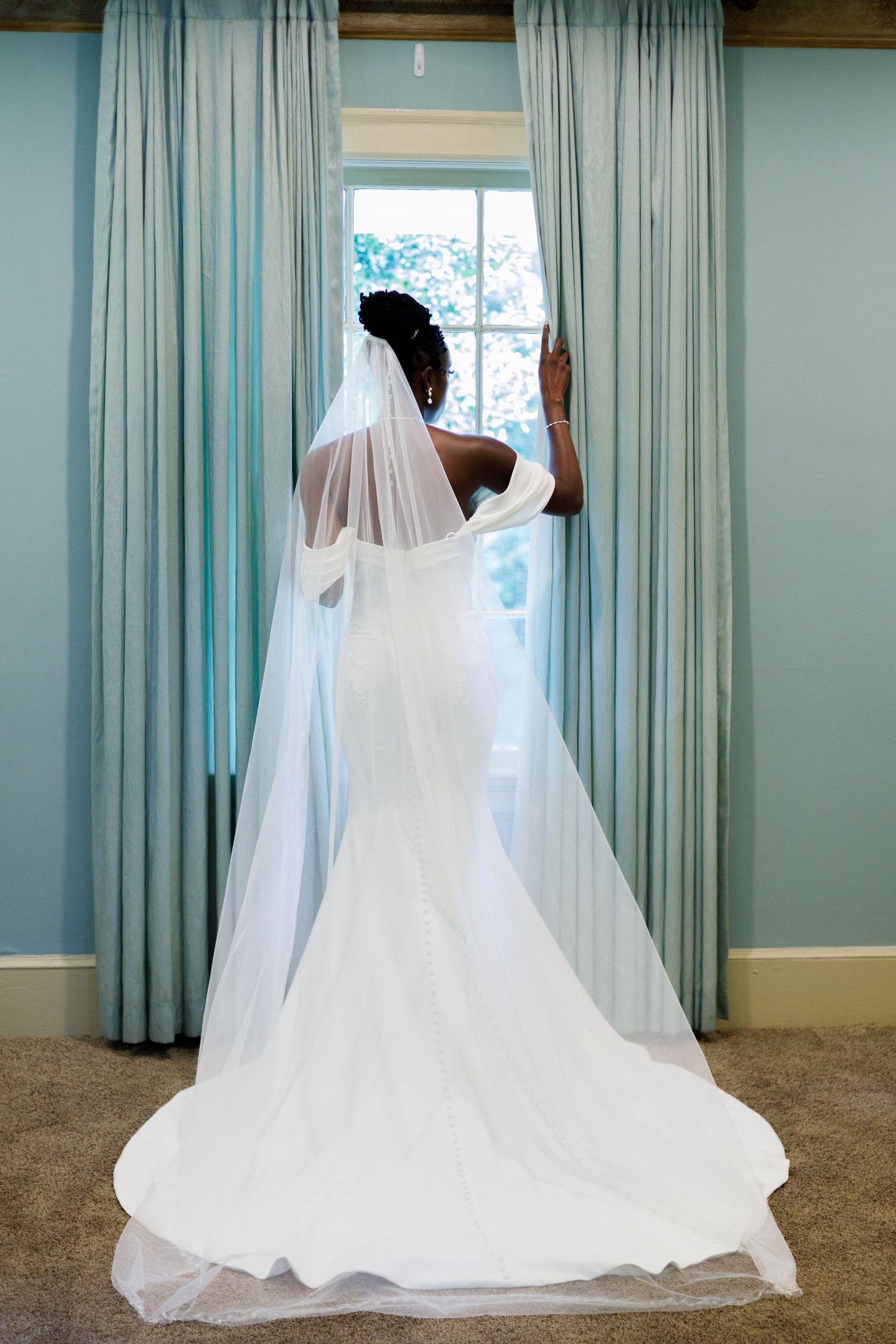 Flowing white wedding dress train and veil create elegant silhouette against blue curtains.