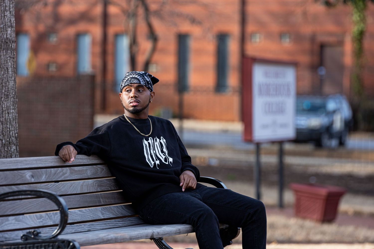 Person in black clothing sits on wooden bench against brick building backdrop in urban setting.