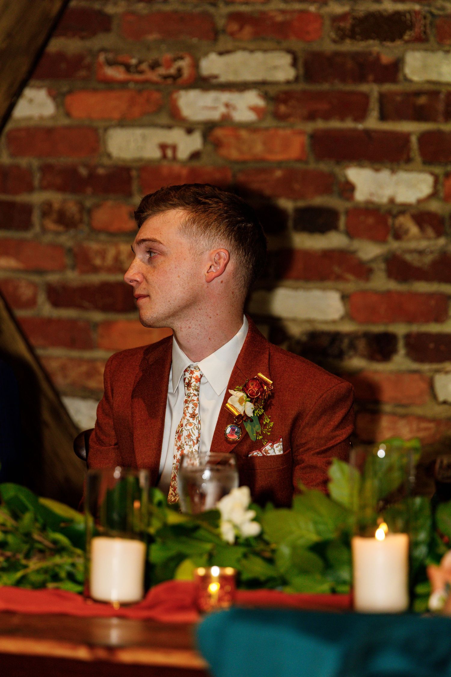 A guest in a burgundy suit jacket sits at an elegantly decorated wedding reception table with candles.