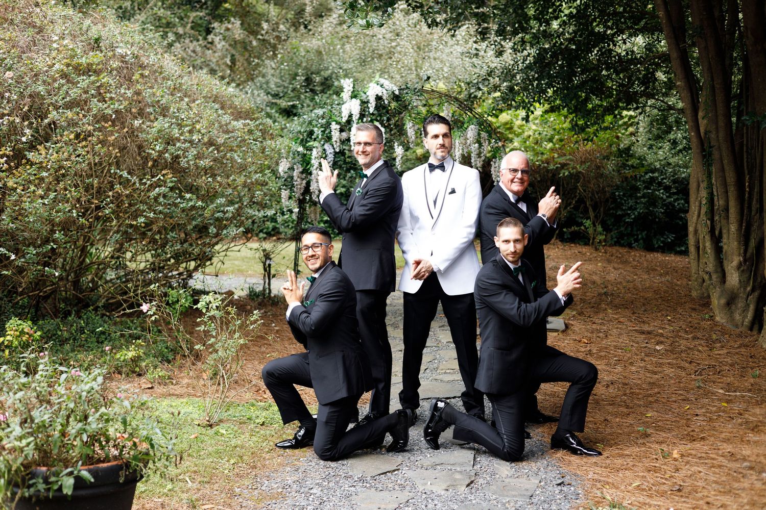 Group of people in formal attire posing playfully in sequence on a wooded path during a photo session.
