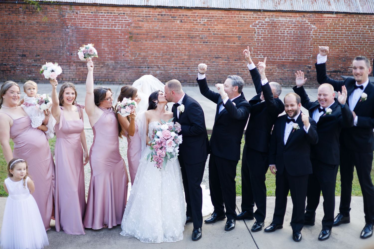 Wedding party lineup with bridesmaids in pink dresses and groomsmen in black suits against brick wall.