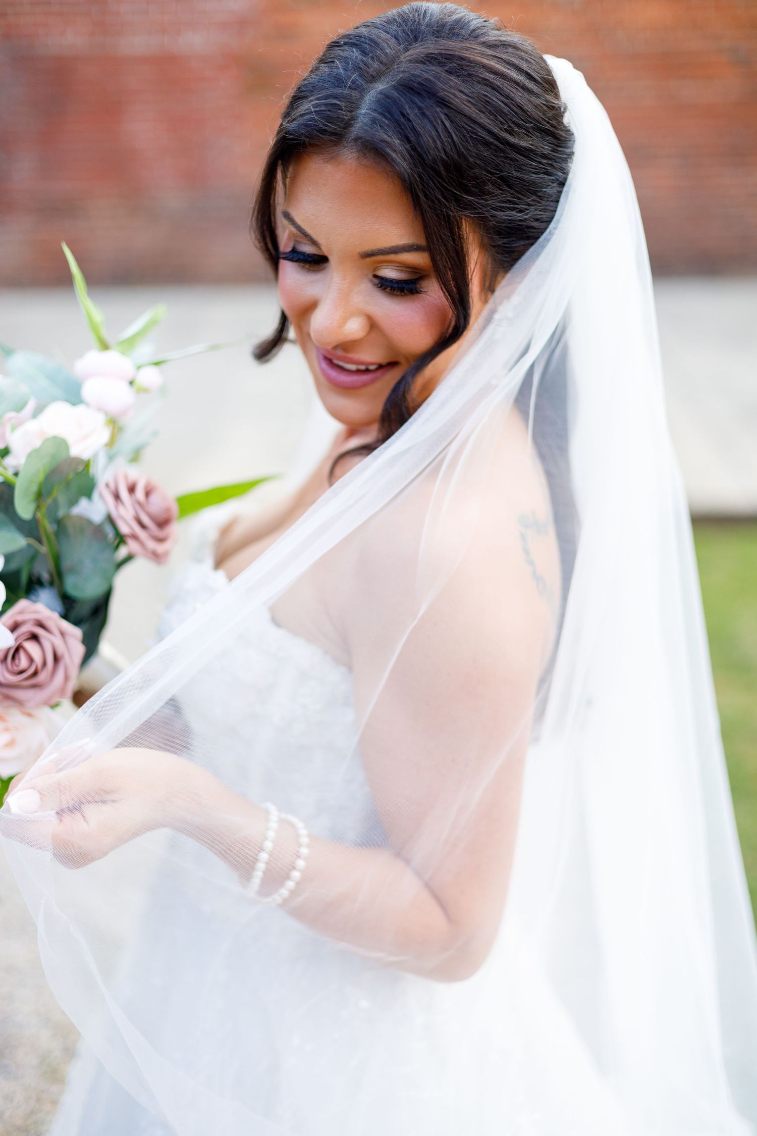 Elegant bride in white gown and flowing veil poses with soft pink roses.