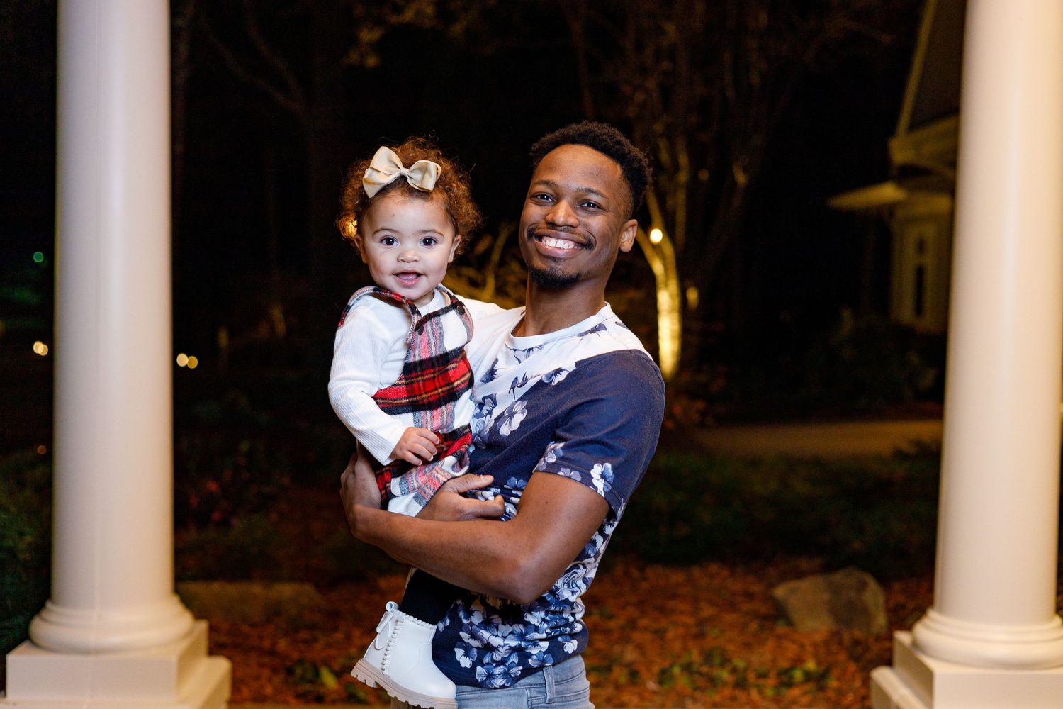A parent and child share a joyful moment together while standing between white columns outdoors at night.