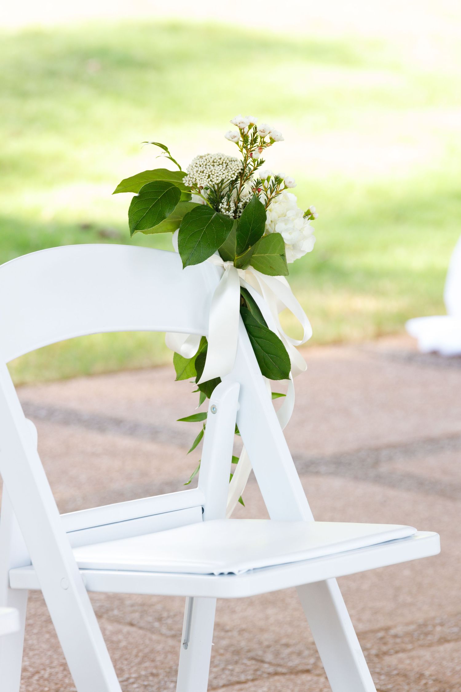 White folding chair decorated with fresh greenery and white ribbon for an outdoor ceremony.