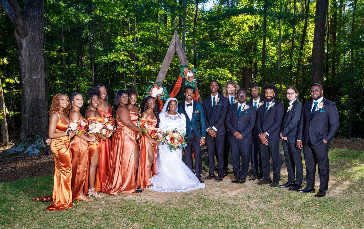 Wedding party members in orange bridesmaid dresses and navy suits gather for formal photos in a forest setting.
