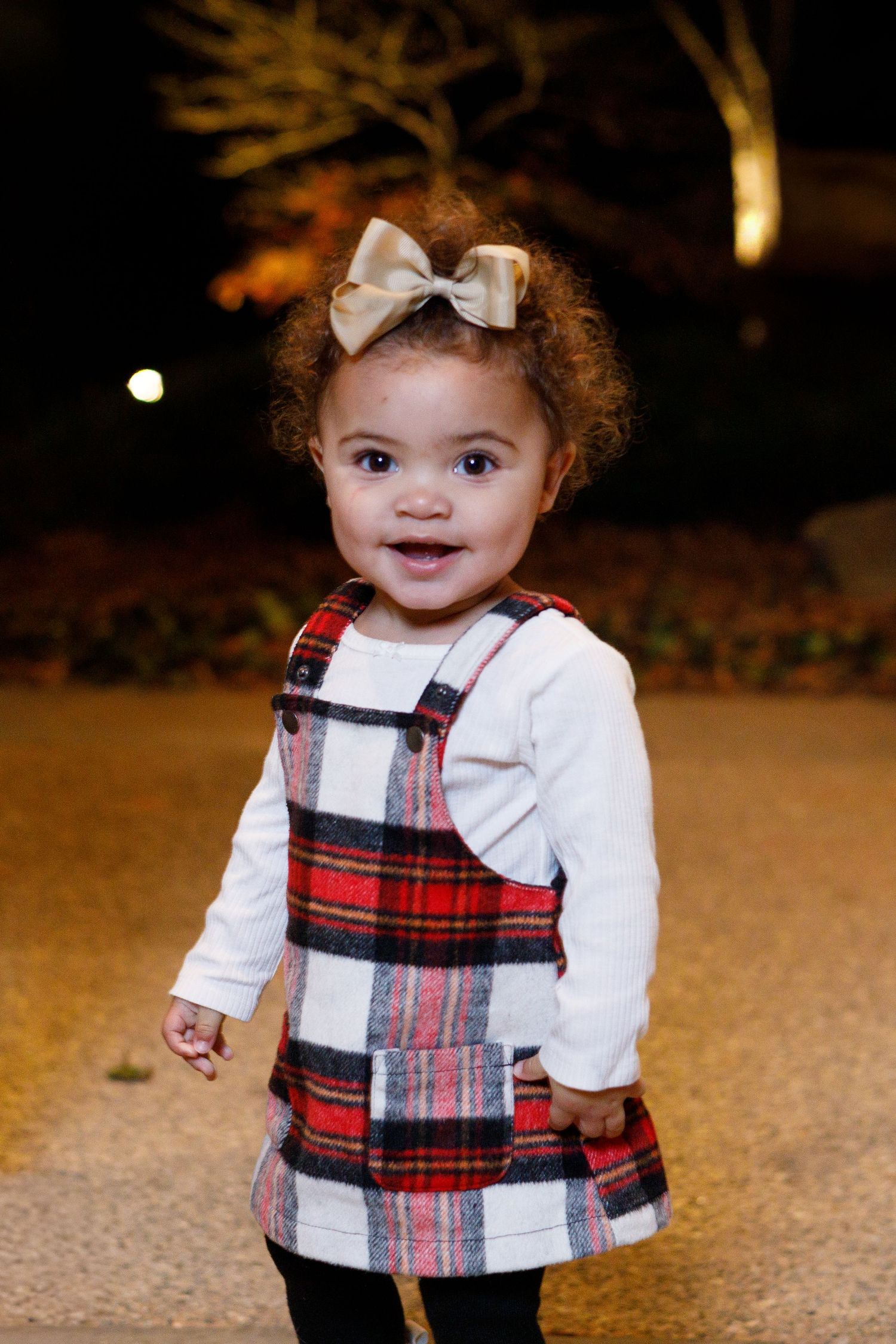 A small child wearing a festive plaid dress with a white bow in their hair smiles at the camera.