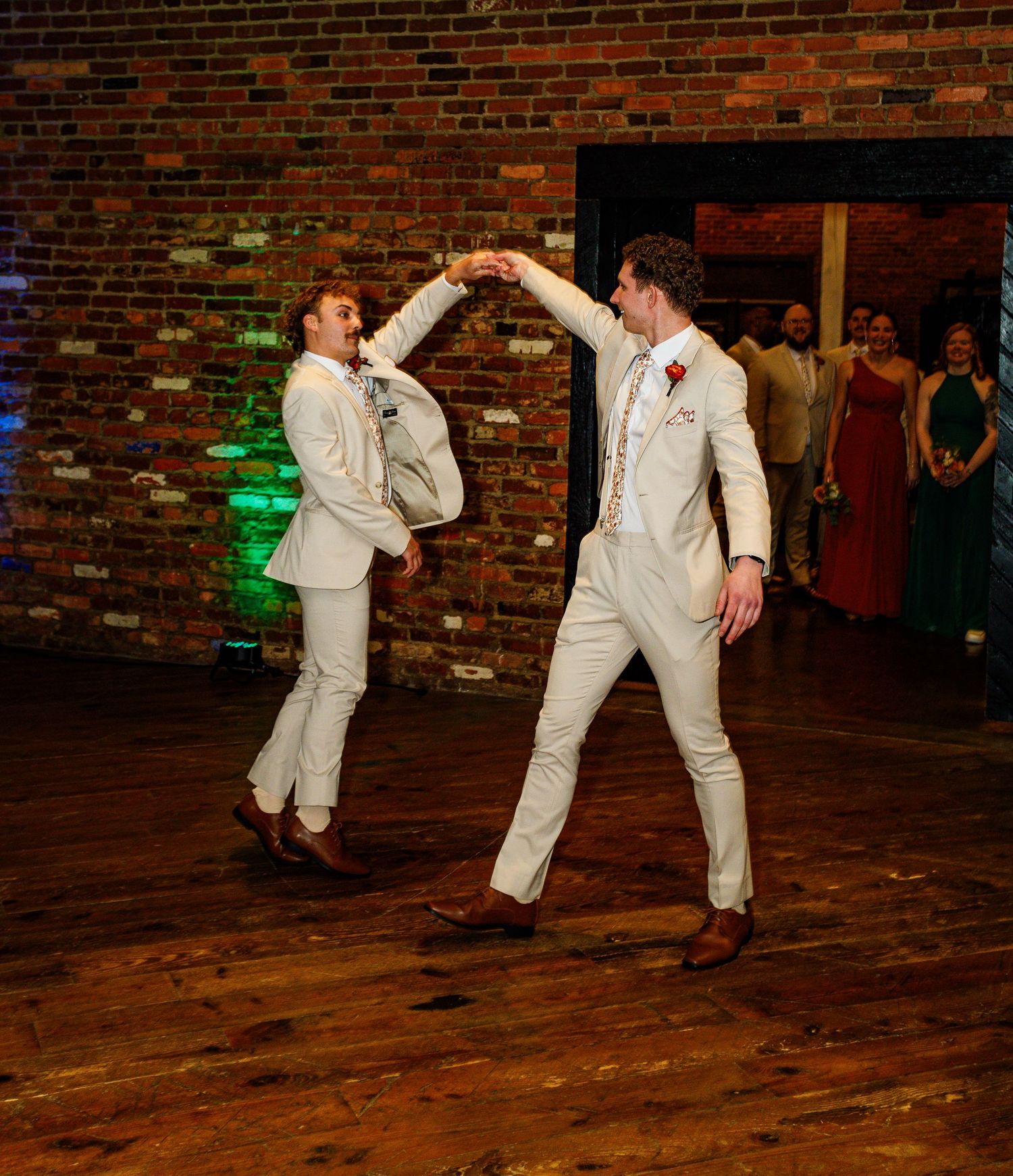 Two people in matching white suits dancing together on a wooden floor with a brick wall backdrop.