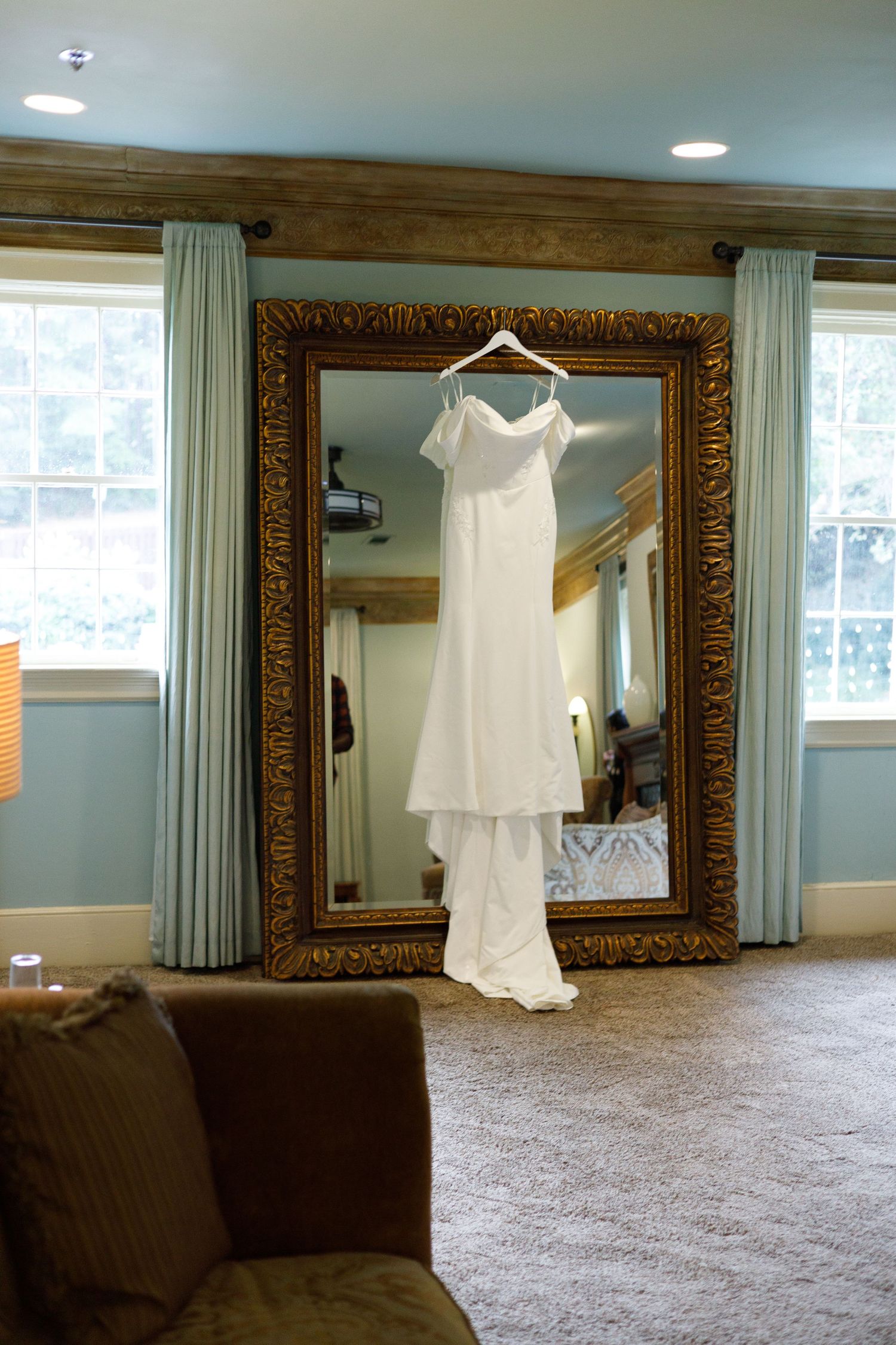 Elegant white wedding dress hanging on display in front of ornate gold-framed mirror.