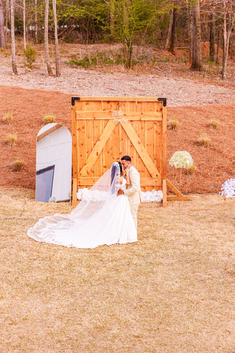 Bride and groom share a romantic kiss in front of a rustic wooden barn door backdrop at an outdoor wedding ceremony.