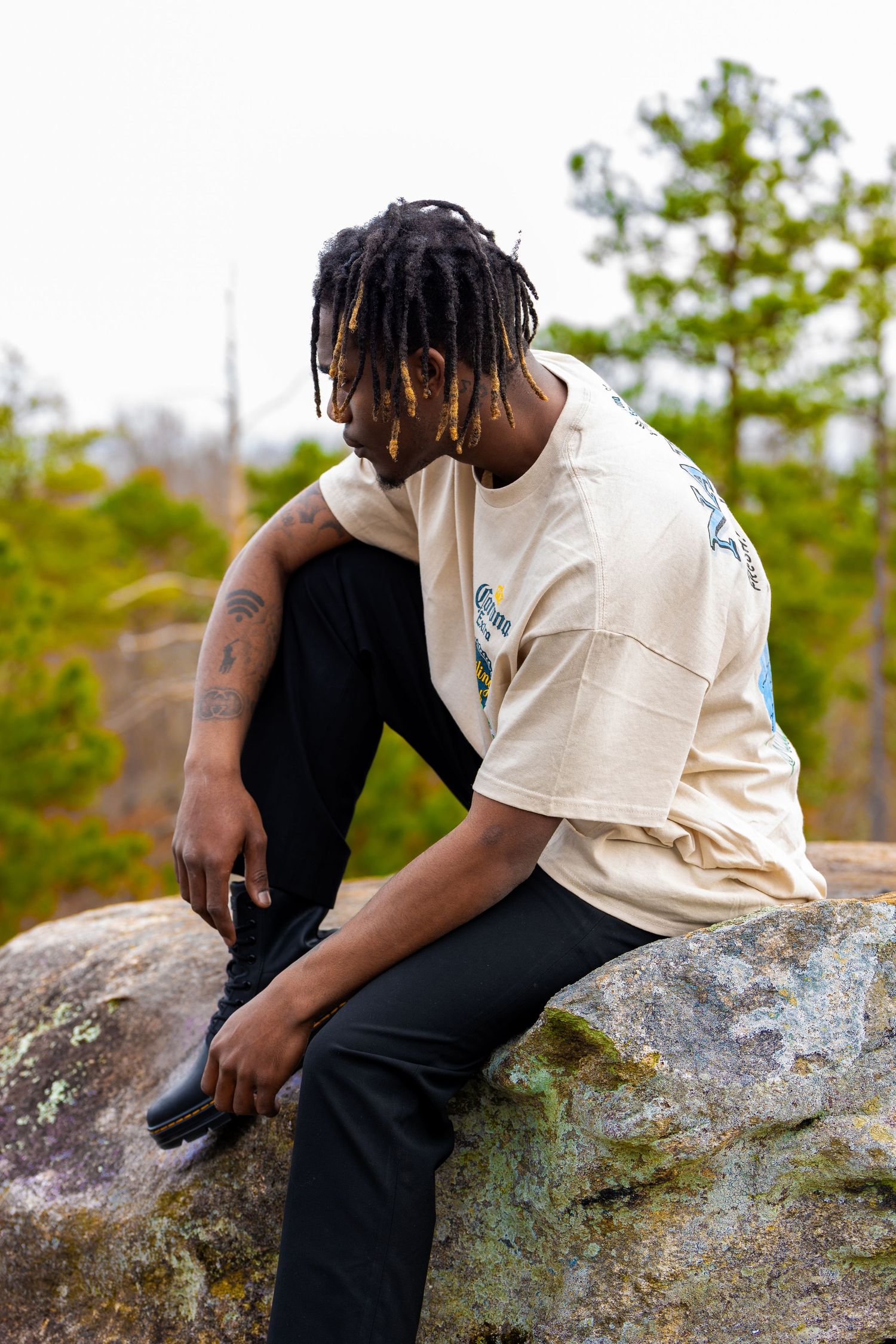 Person in black and cream outfit sits thoughtfully on large boulder in natural outdoor setting.