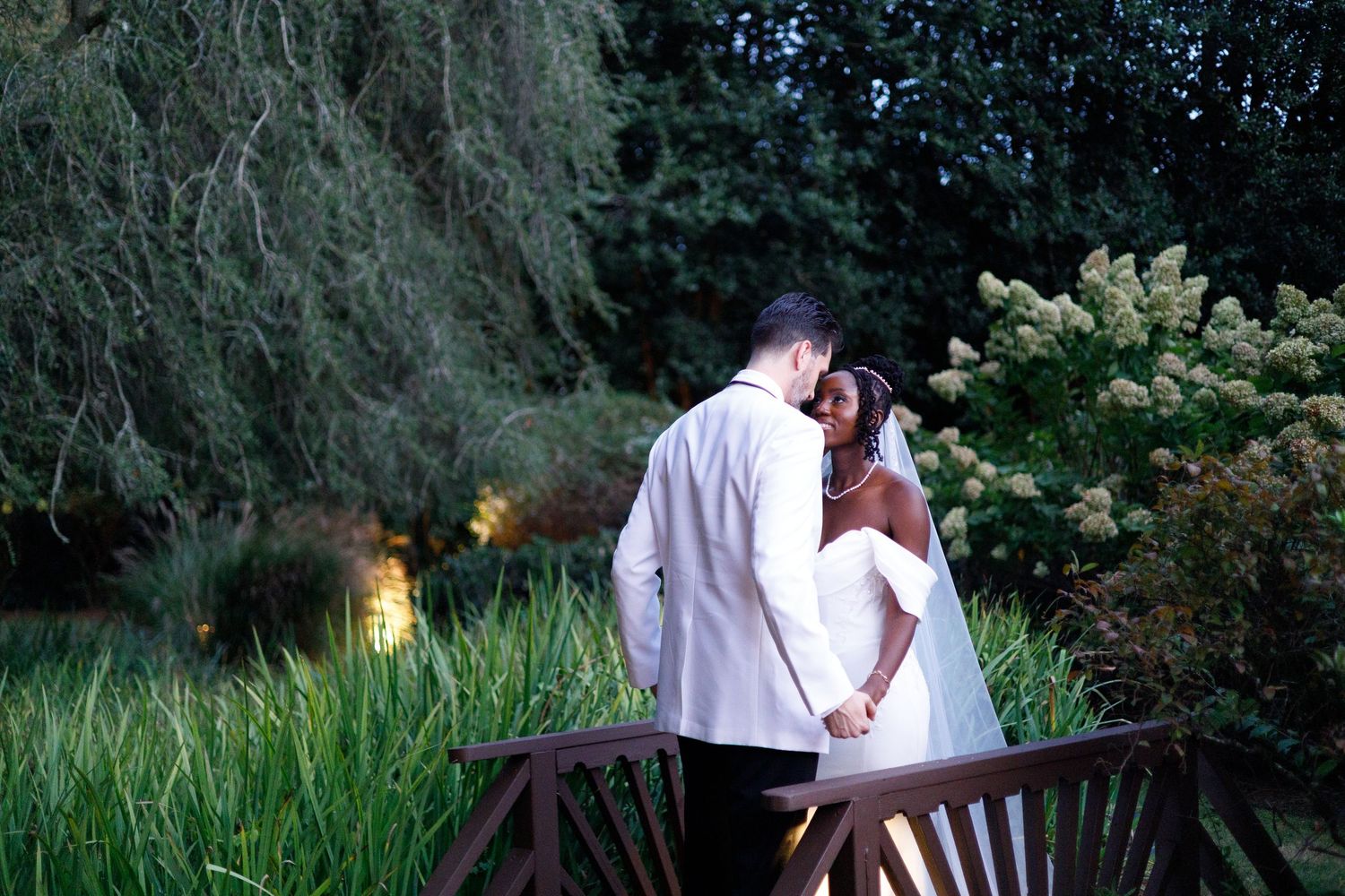A romantic couple sits together on a wooden bench in a garden setting at dusk.