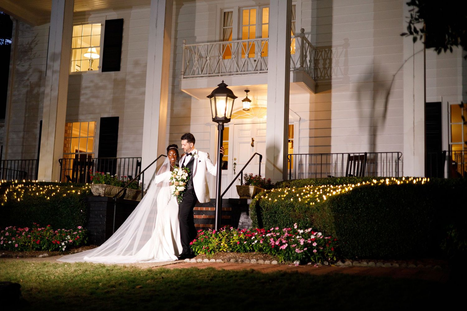 Wedding couple silhouetted against illuminated white mansion at night.