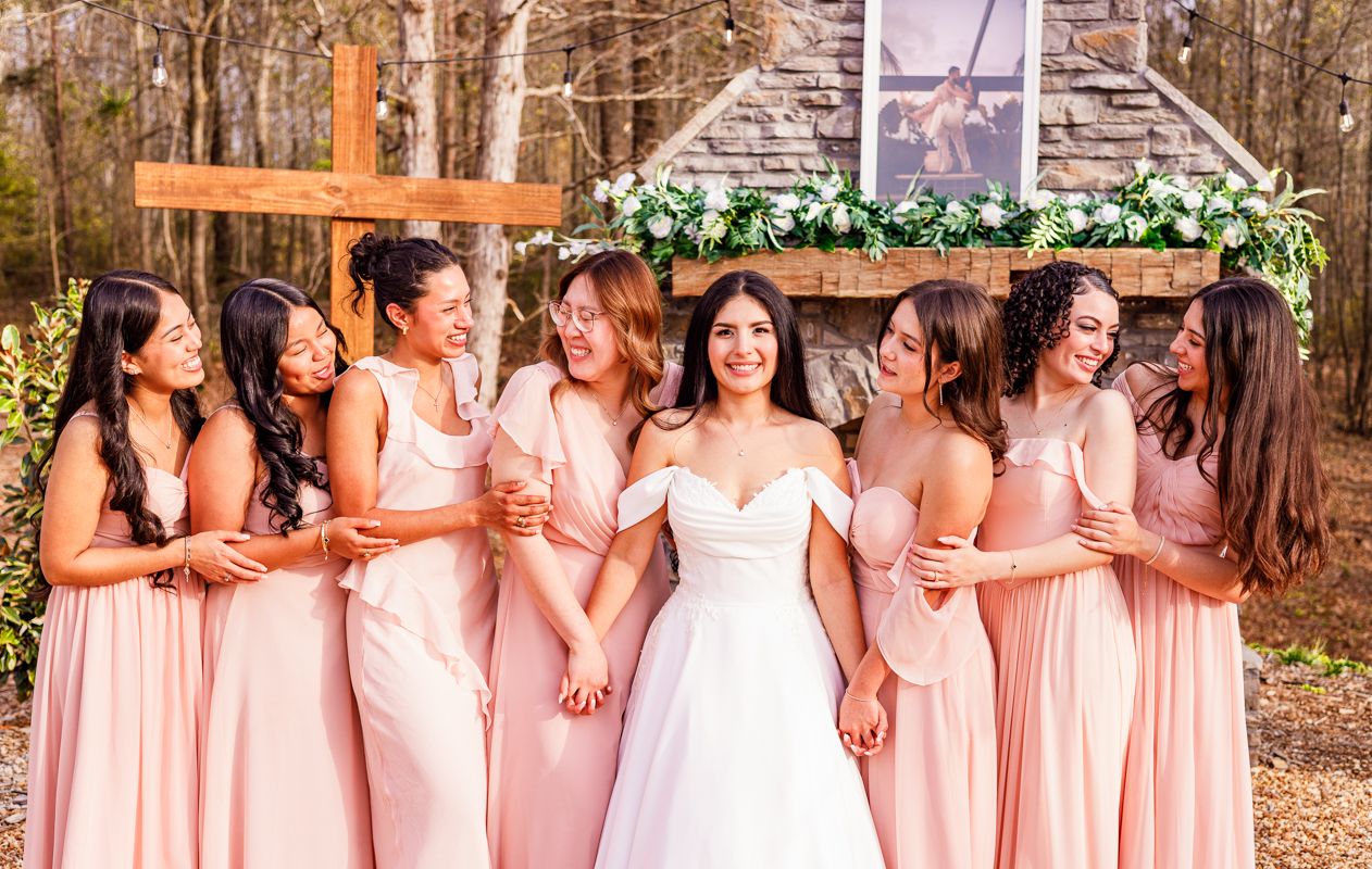 Bride in white gown surrounded by six bridesmaids in blush pink dresses, laughing outdoors near a floral arch.