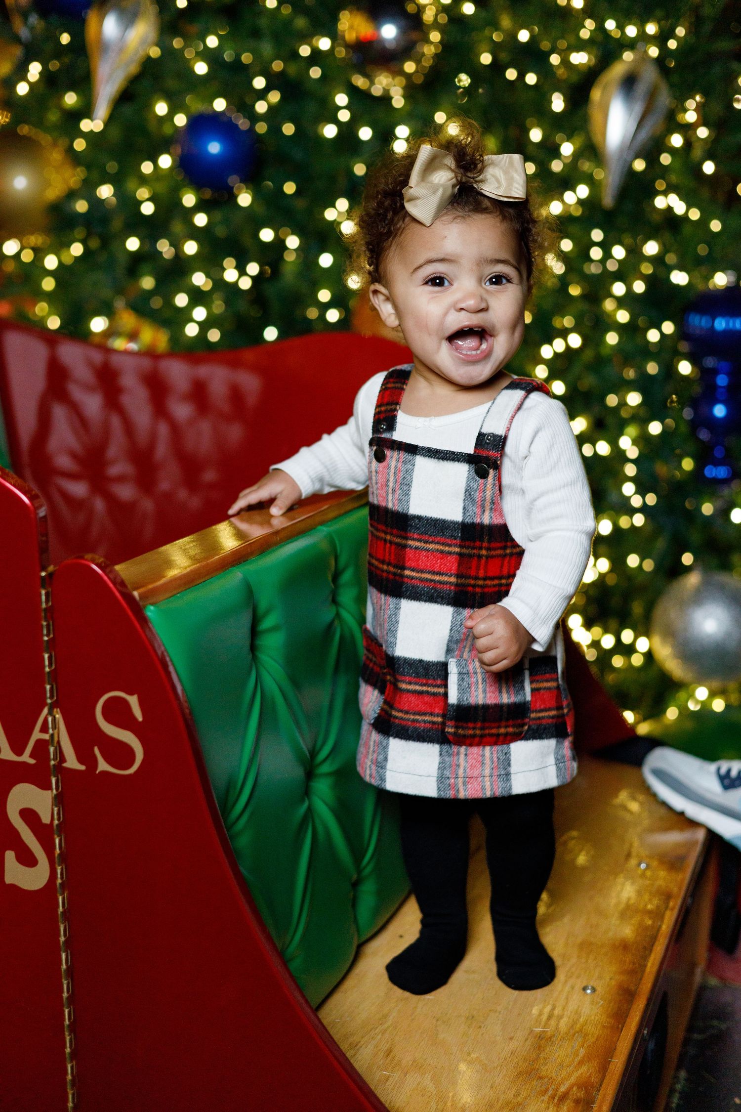 Festive holiday photo with plaid dress and bow against Christmas tree lights and decorative red sleigh backdrop.