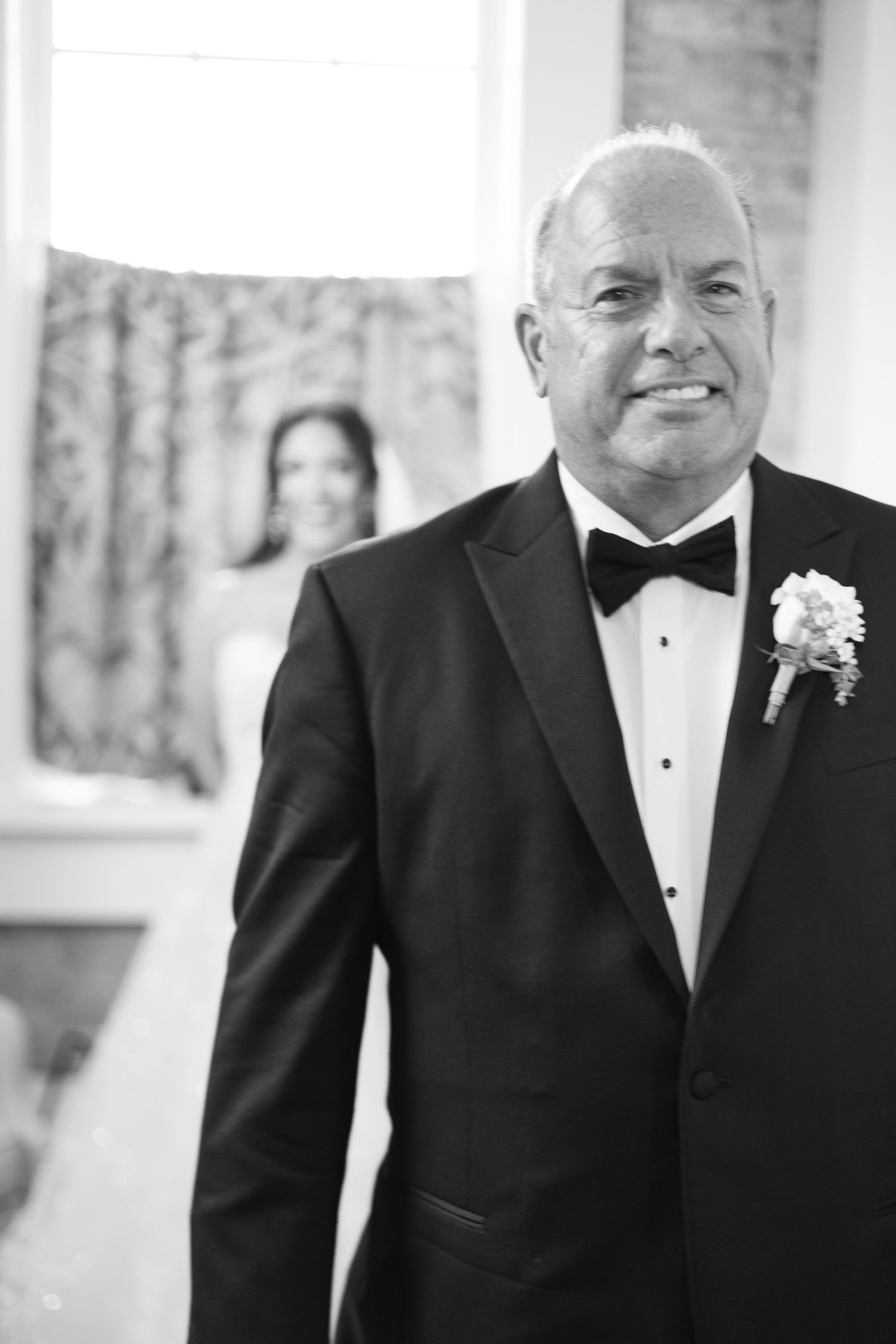 A formal portrait of a person wearing a classic black tuxedo with a white boutonniere.