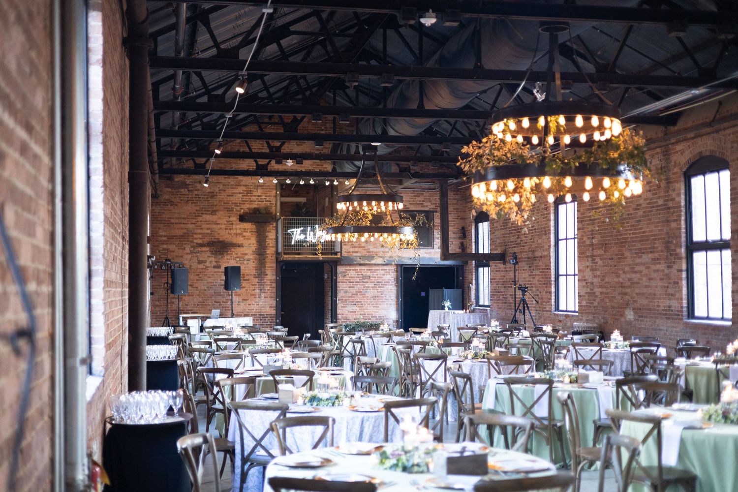 Rustic wedding reception venue with exposed brick walls, wooden beams, and elegant chandeliers illuminating dining tables.