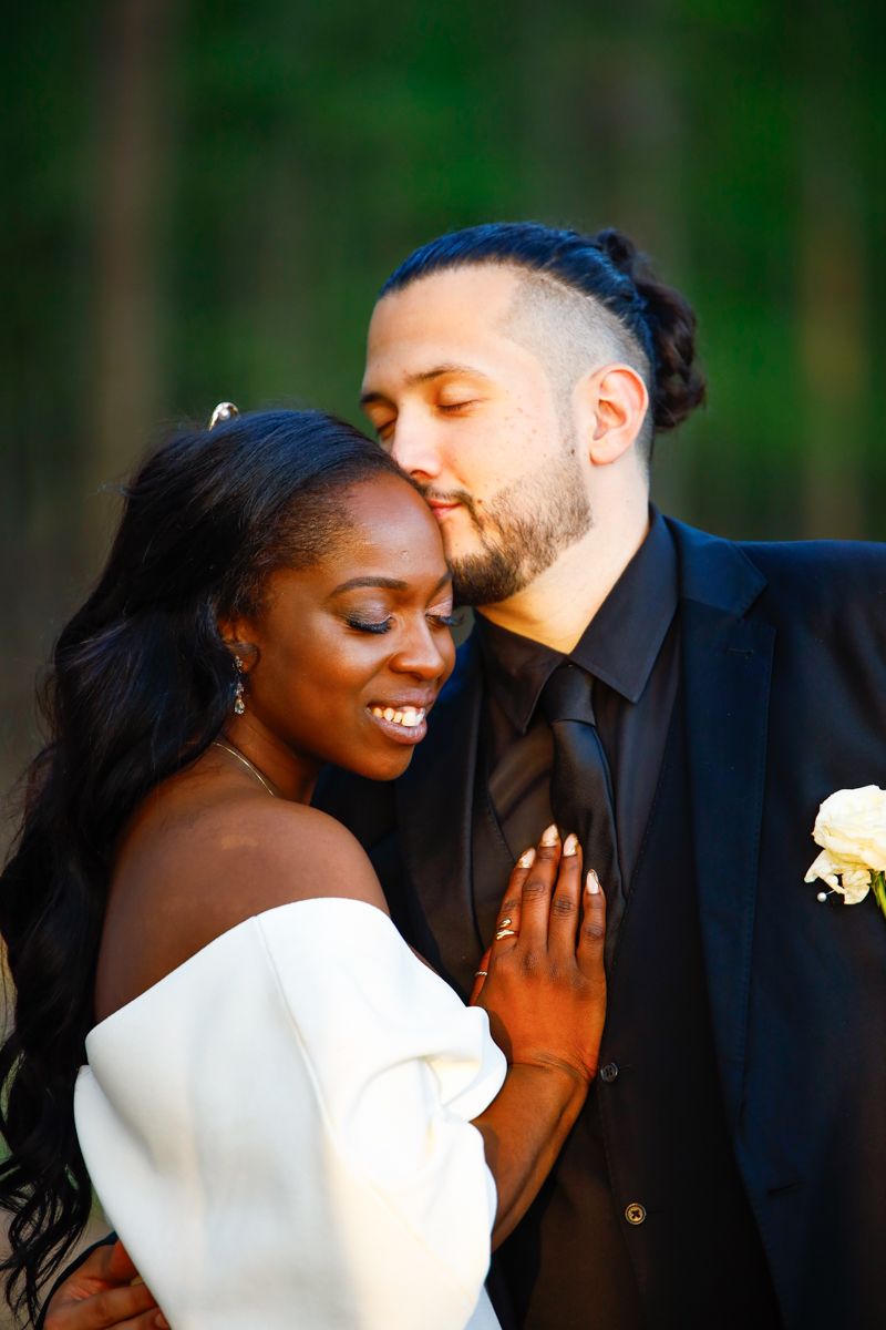 A tender wedding moment captured outdoors with a bride in white off-shoulder dress and groom in black suit.