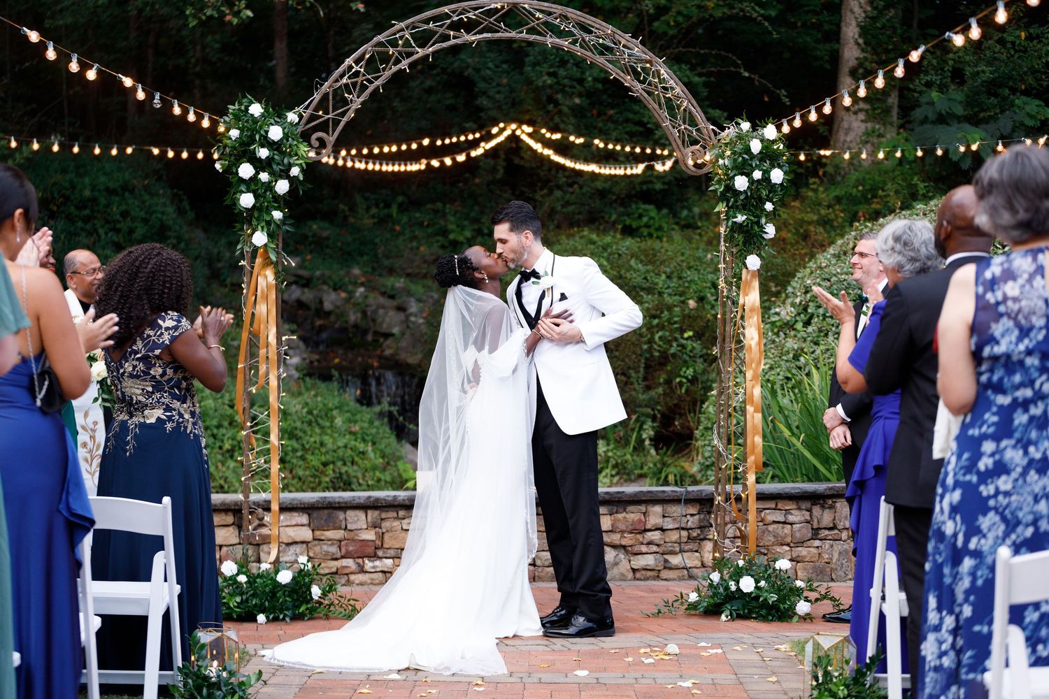 Bride and groom share first kiss under floral arch with string lights at outdoor wedding ceremony.