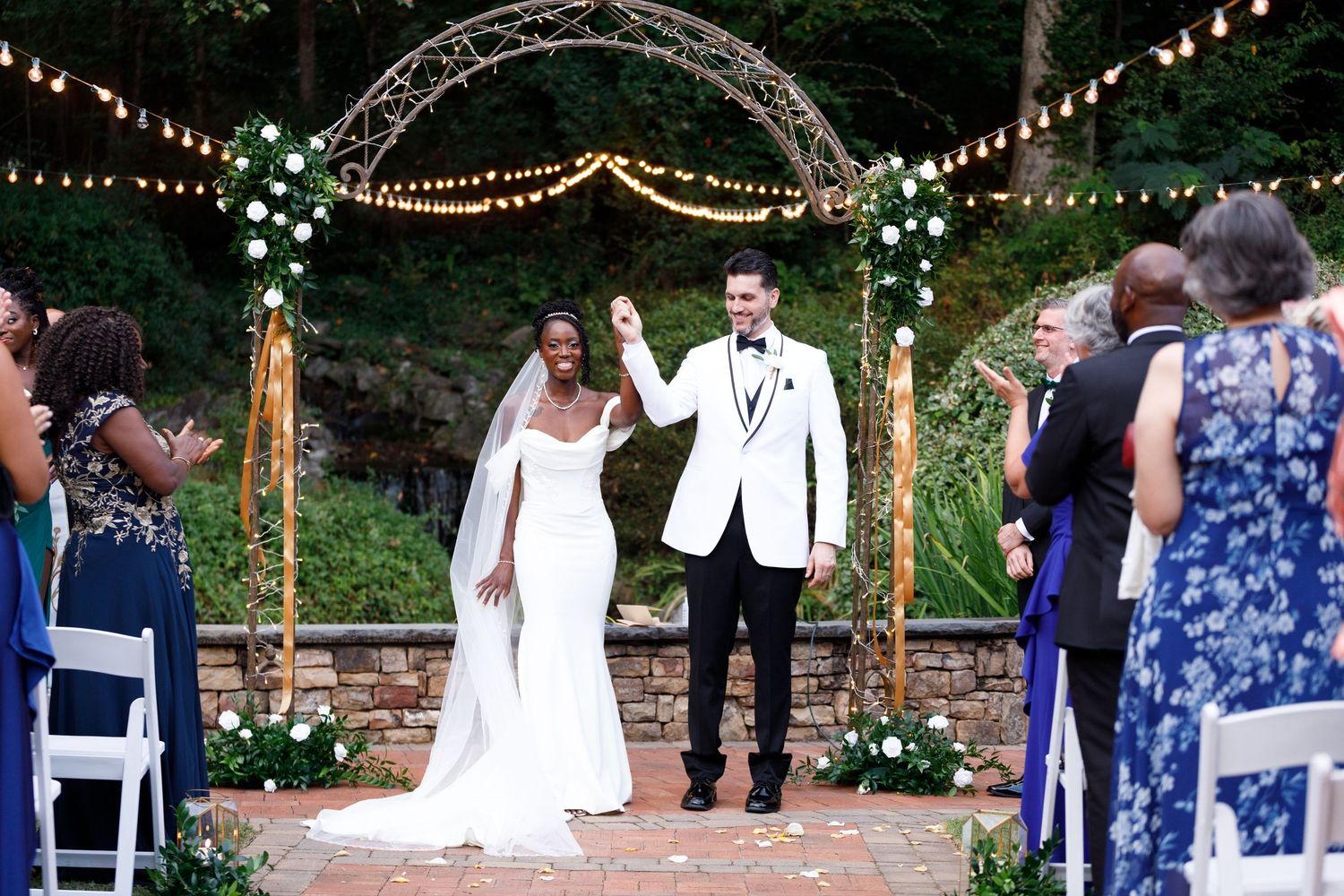 Outdoor wedding ceremony setup with string lights and white chairs under evening sky.