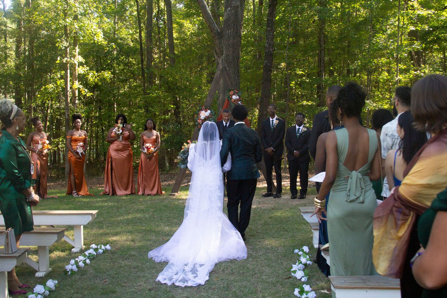 Outdoor forest wedding ceremony with bridal party in coral dresses and guests watching.