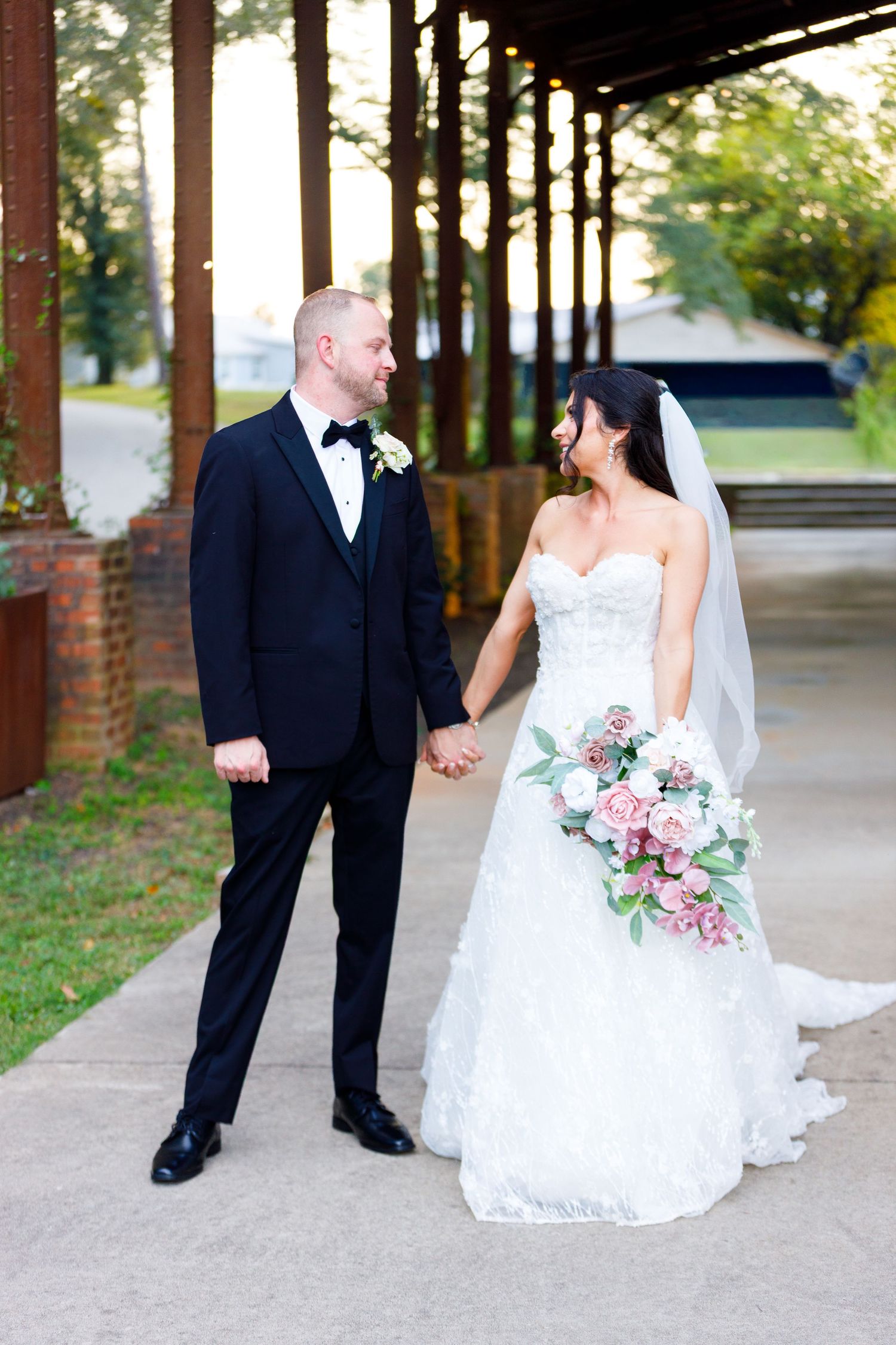 Newlyweds share romantic moments under covered brick walkway with pink floral bouquet.