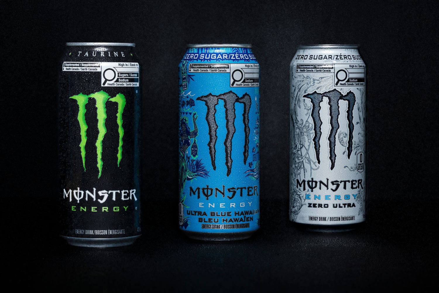 Three Monster Energy cans (different flavors) lined up on dark reflective surface.