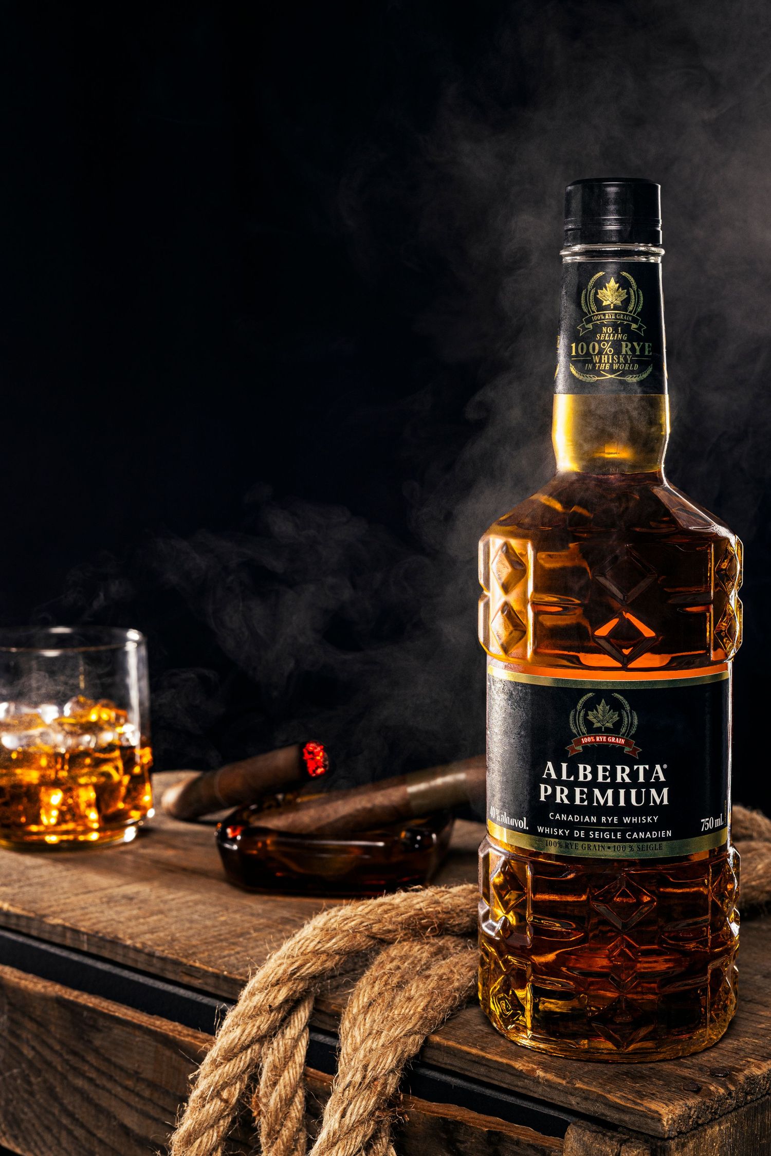 Dramatic product photography of Alberta Premium whisky bottle with cigar, glass, rope, and smoke on wooden table.