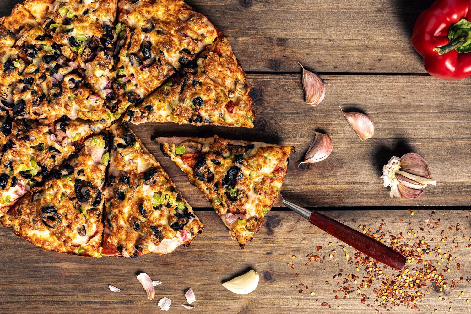 Close-up food photography of sliced loaded pizza with olives, peppers, and cheese on wooden table.