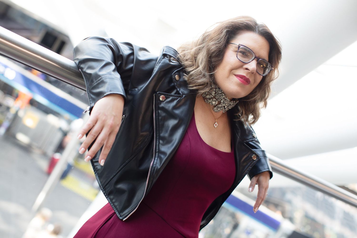 Woman in black leather jacket and burgundy dress posing outdoors wearing glasses and statement necklace.
