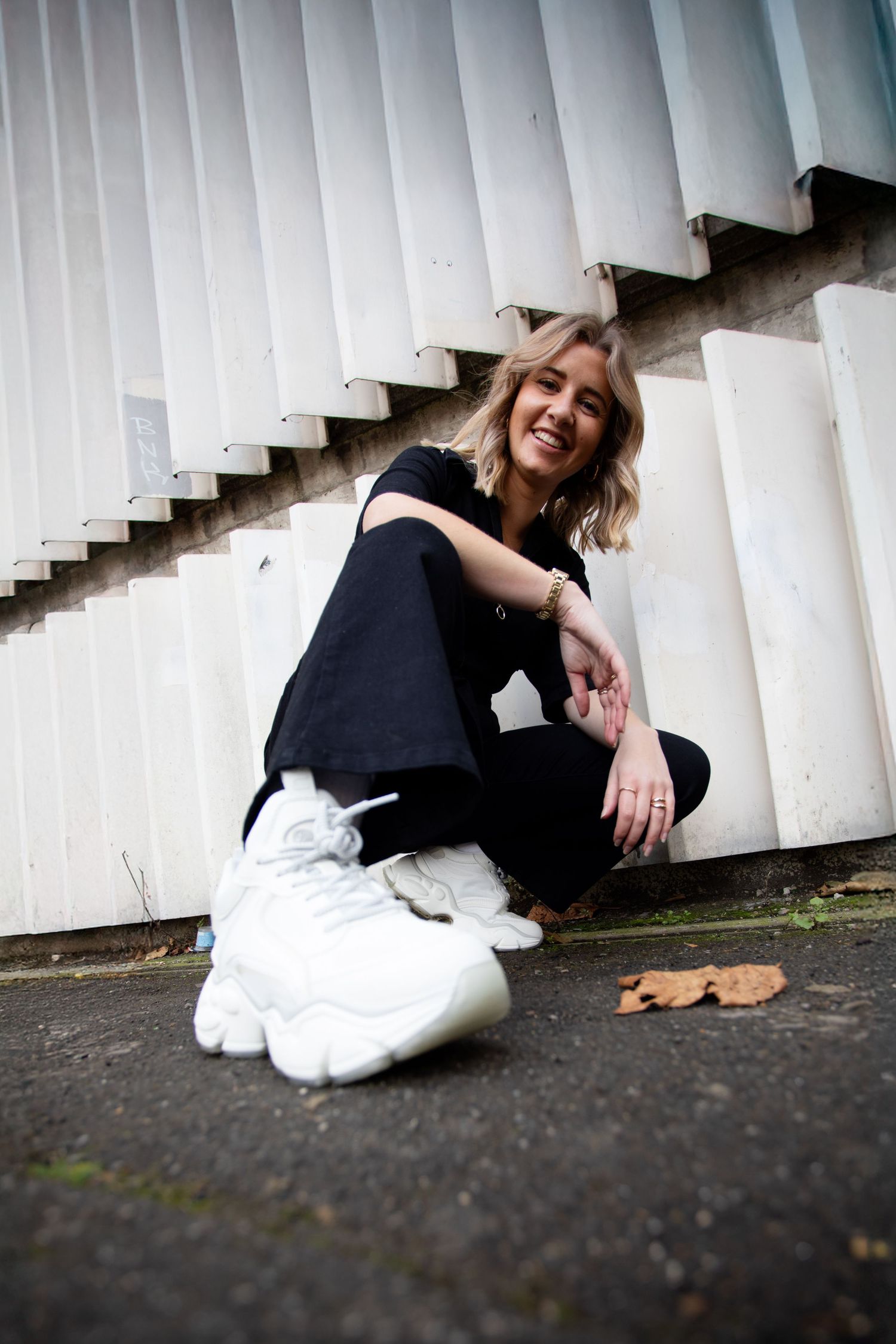 Smiling blonde woman in black outfit and white sneakers sitting cross-legged outdoors against corrugated metal wall.