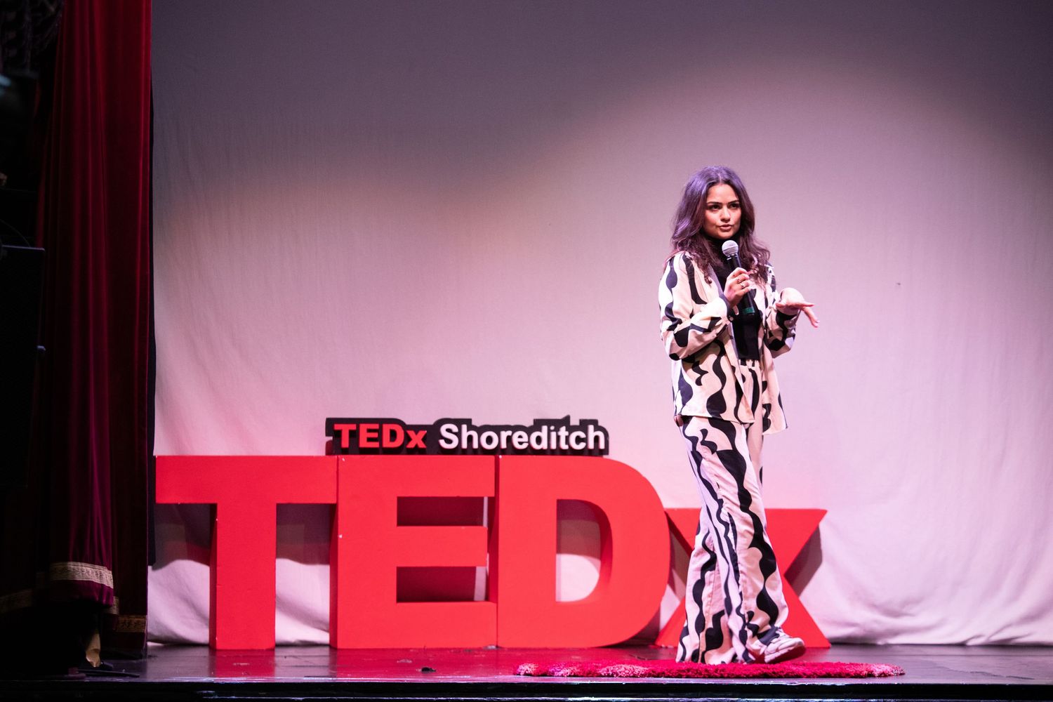 A woman in a patterned gown speaks on stage at TEDx Shoreditch event with bold red TEDx letters behind her.
