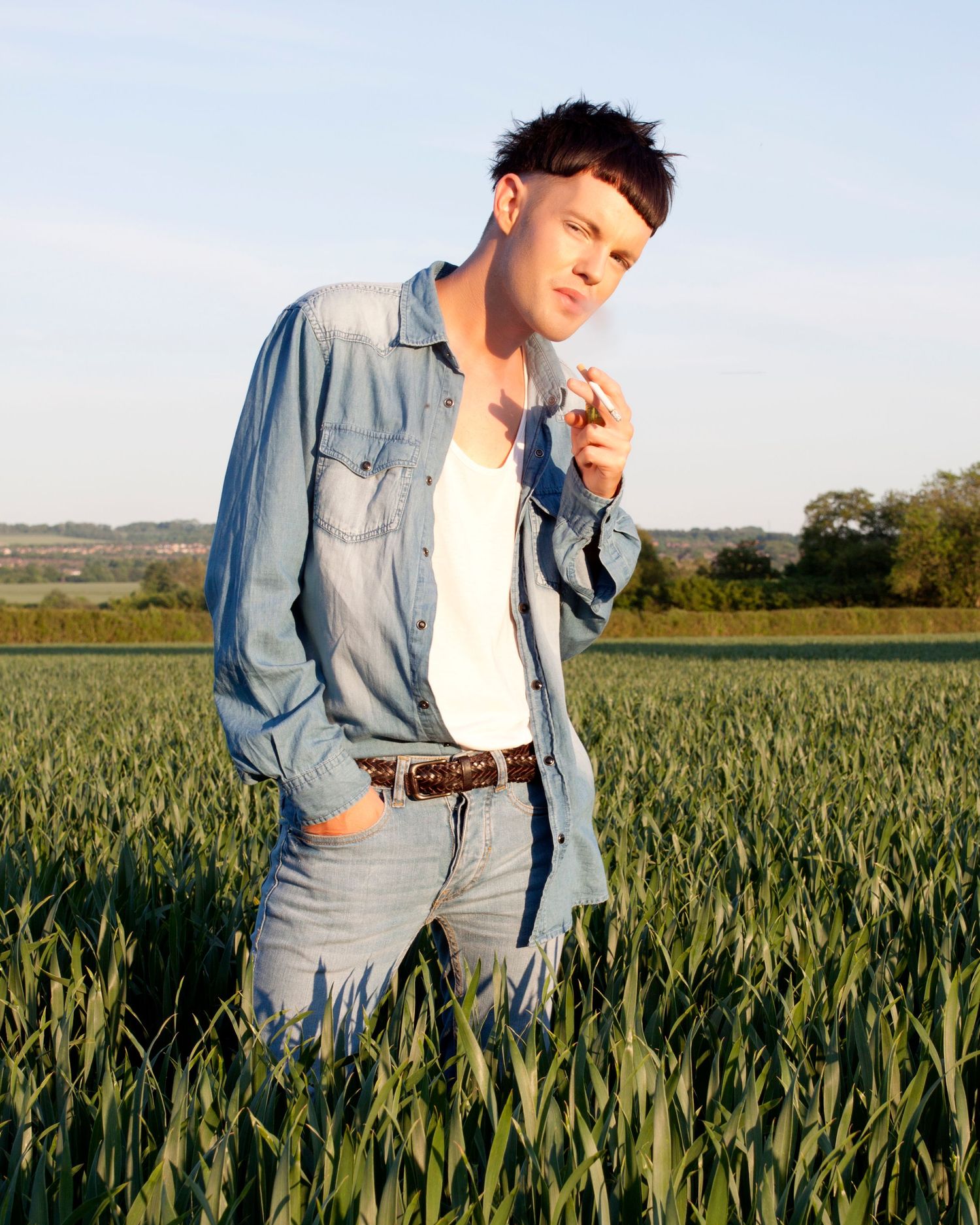 Casual portrait of someone wearing denim clothing standing in a green wheat field at sunset.