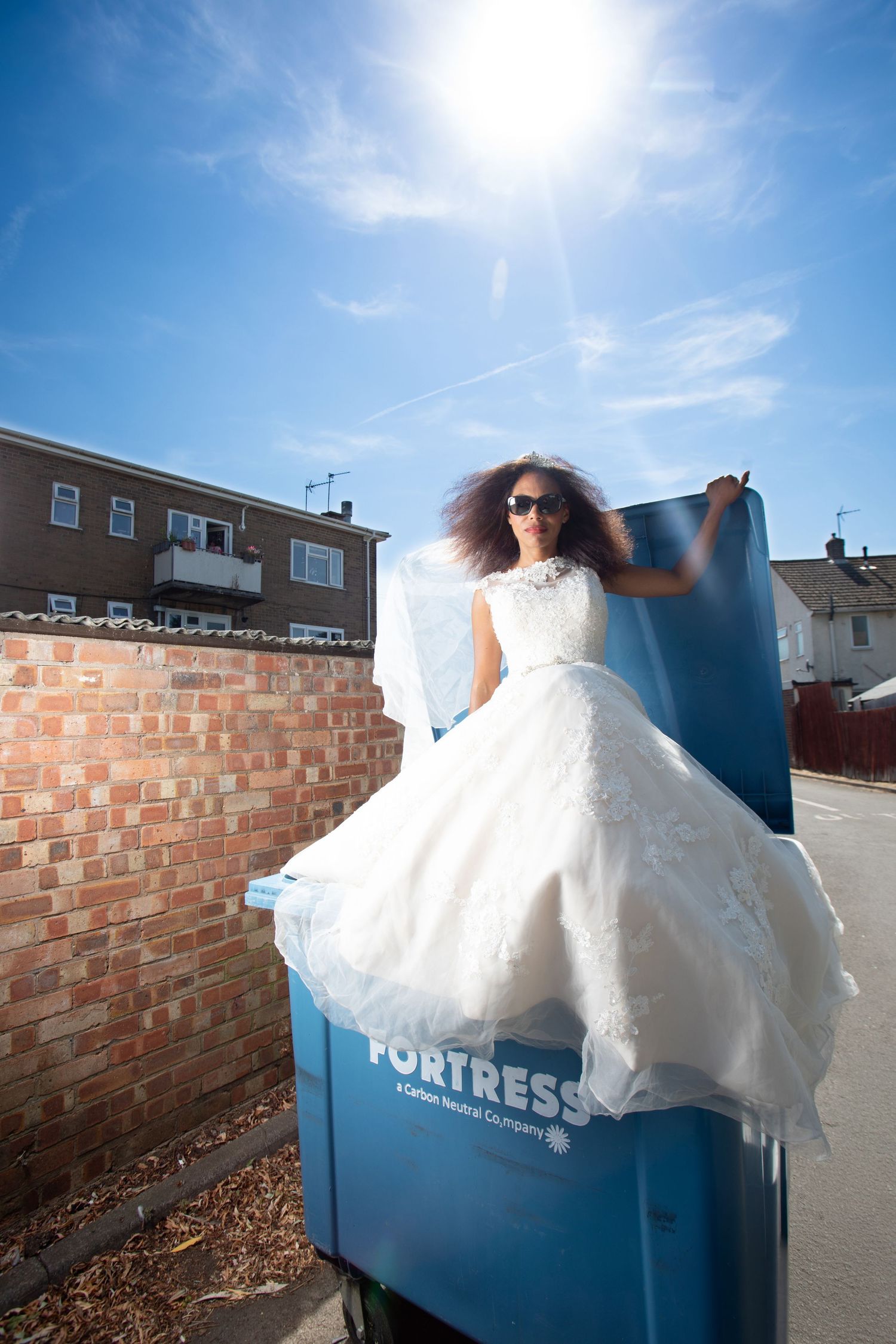 Woman in white wedding dress posing on a blue Fortress dumpster outdoors on a sunny day.