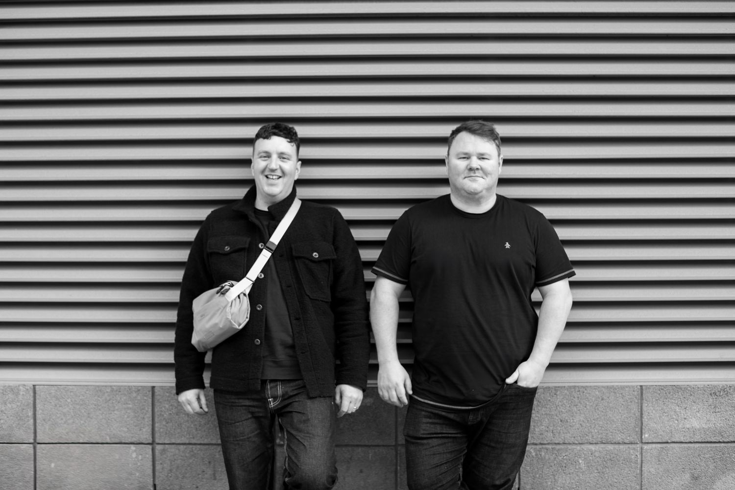 Two men in black clothing pose against a striped wall for a monochromatic portrait.