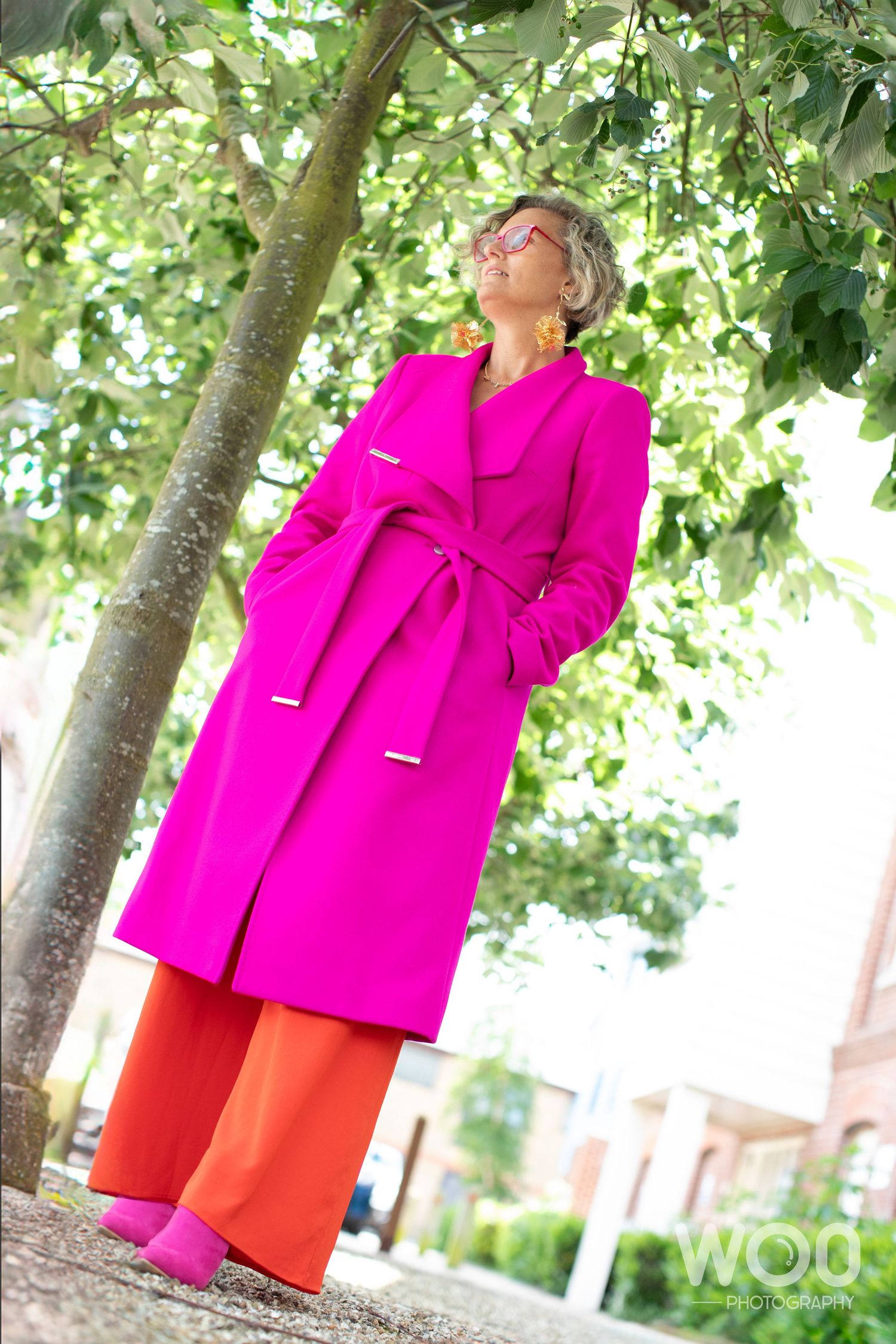 Bold fashion look featuring hot pink coat and orange pants under leafy tree.