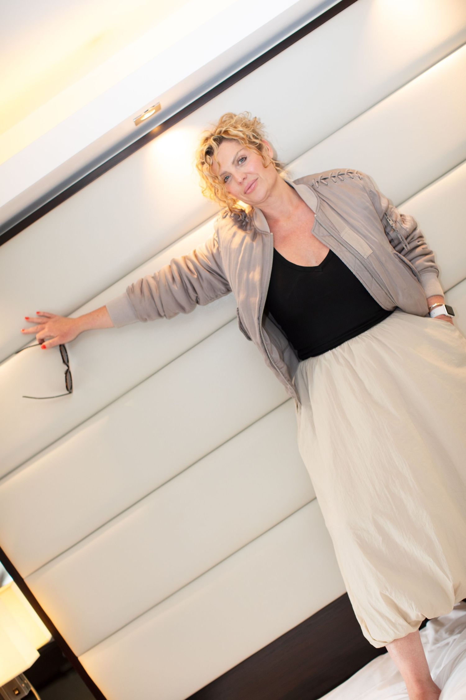 Blonde woman in beige skirt and jacket posing confidently against a white padded wall in a modern interior.