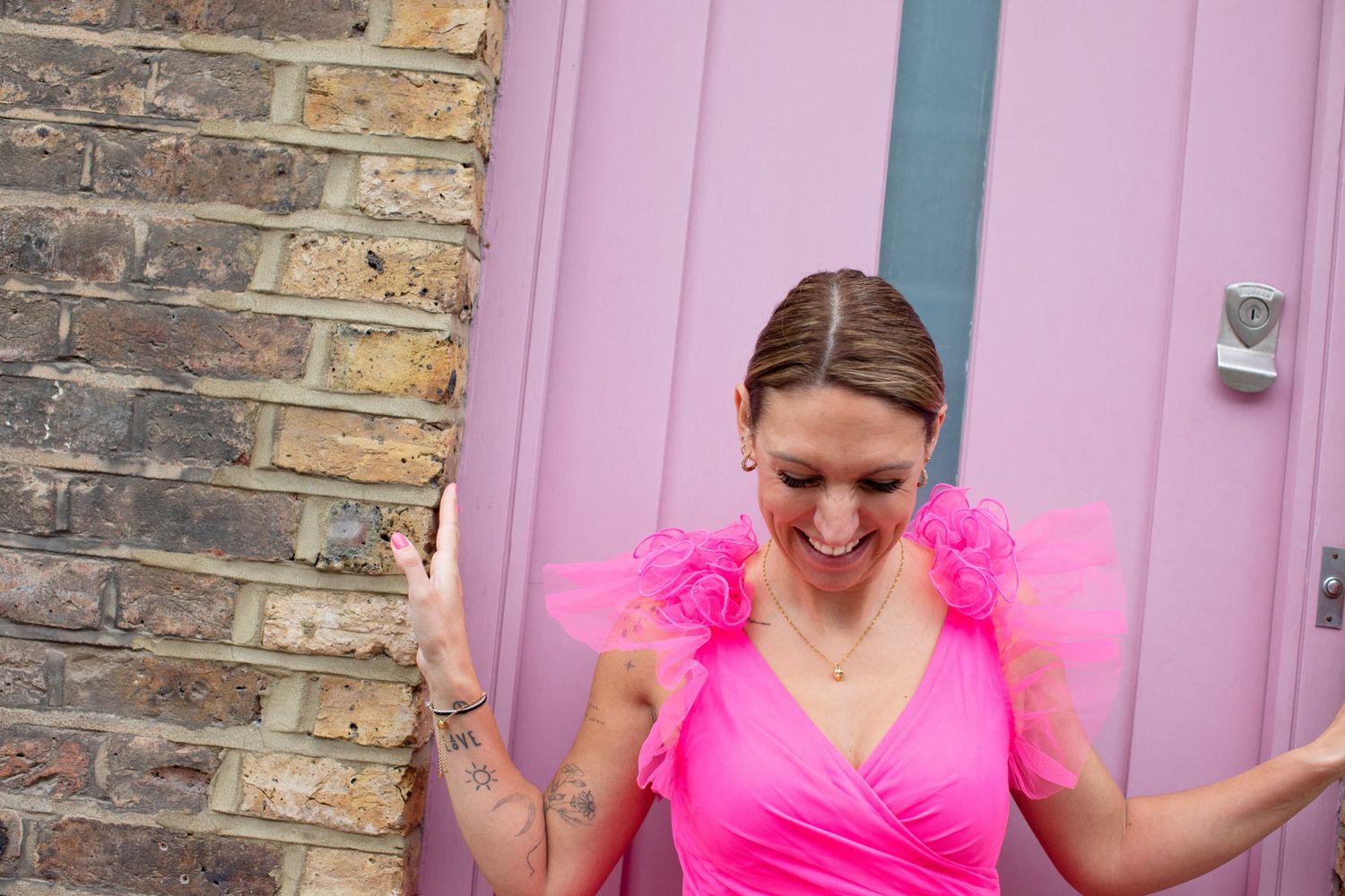 Woman in bright pink ruffle dress laughing joyfully outside a pink door with brick wall background.