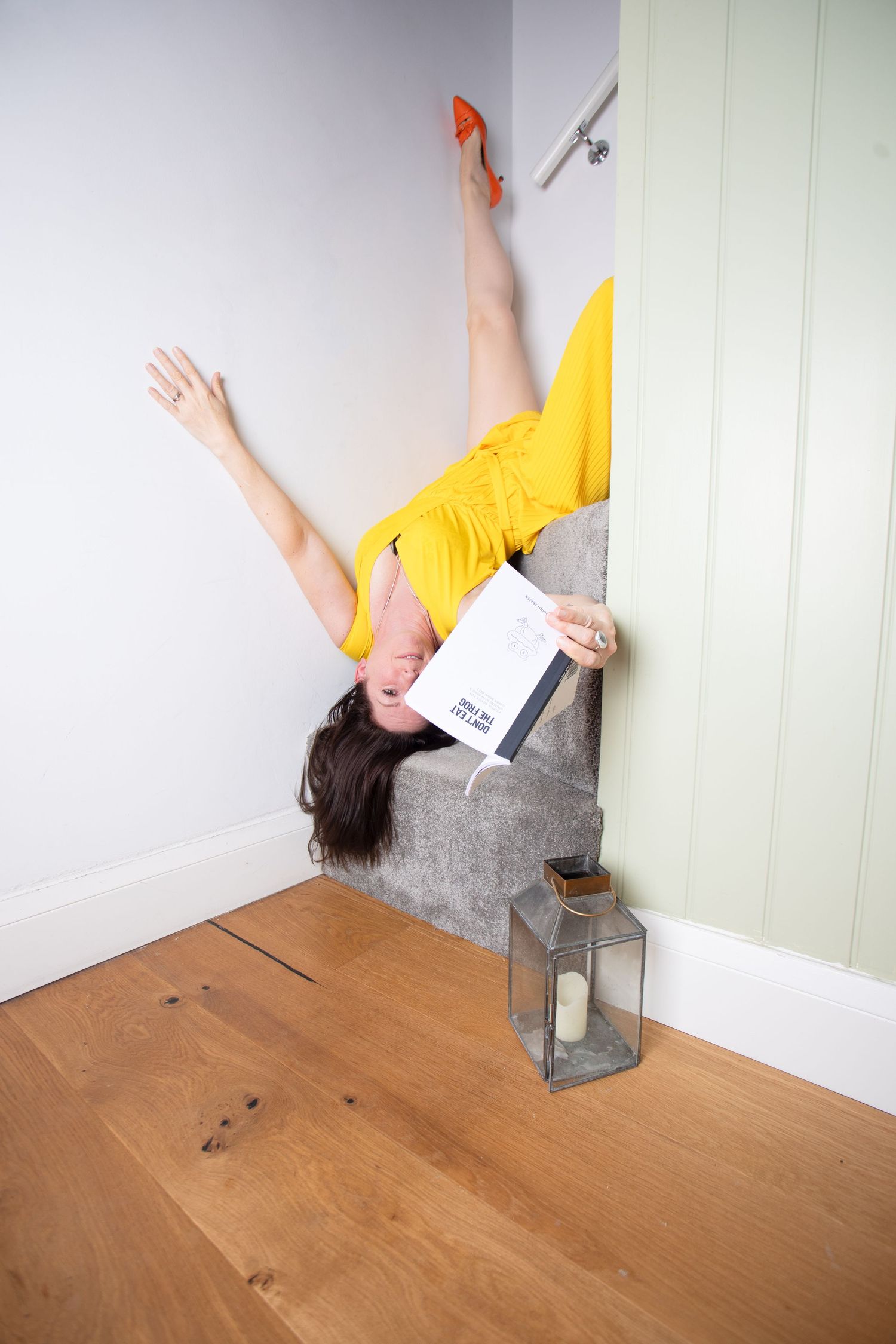Woman in yellow dress and orange heels tumbling upside down over stairs while holding a book indoors.