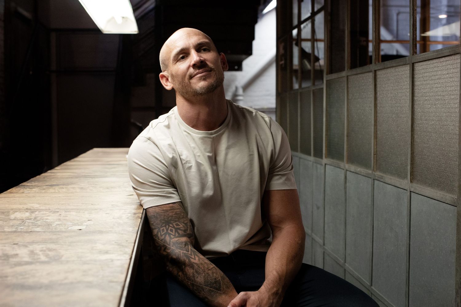 Tattooed bald man in white t-shirt smiling upward while seated at wooden counter in dimly lit space.
