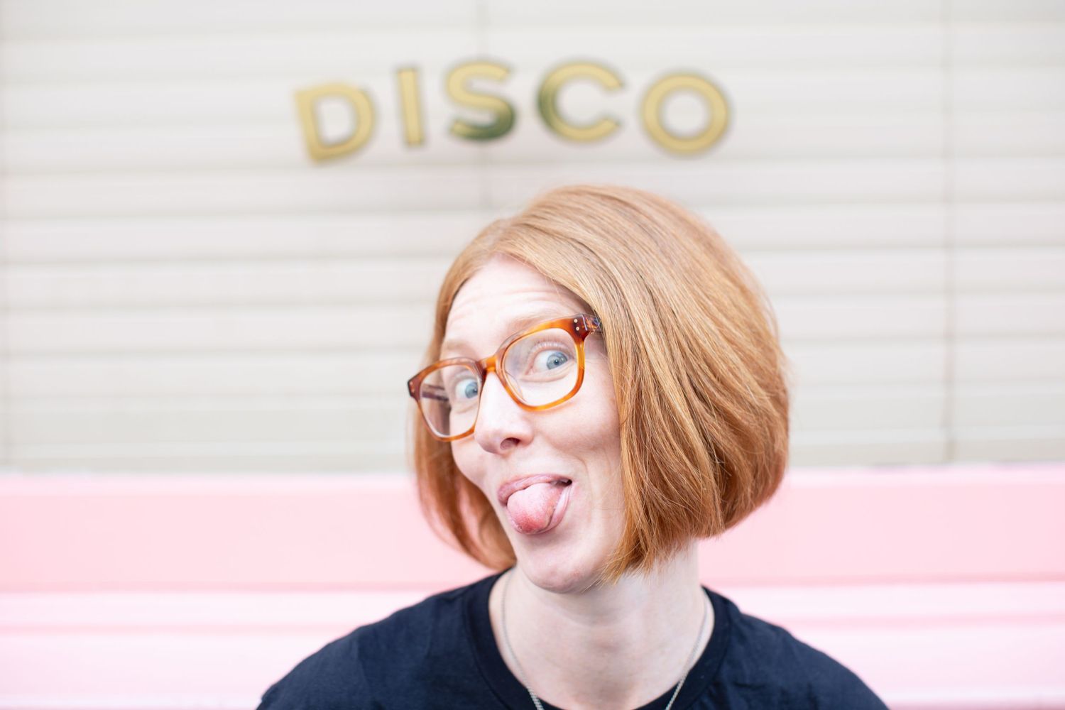 Young redhead woman with round glasses sticking out tongue playfully in front of a pink and white DISCO sign.